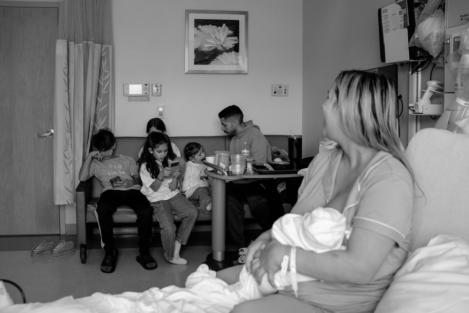 Black and white photo showing a group of people gathered in a room with some sitting at a table while another rests on a bed.