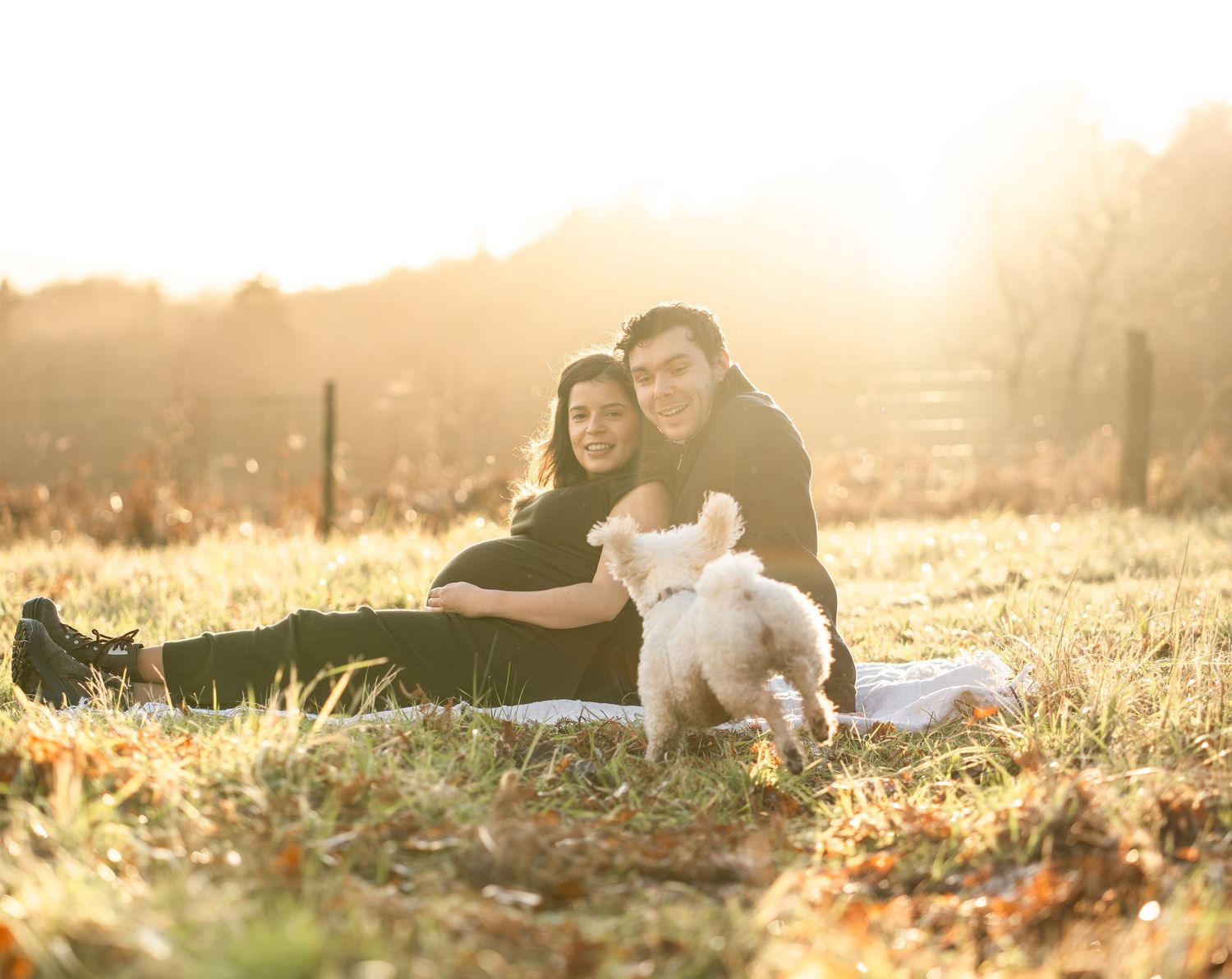 Couple lying together in a sunlit meadow during golden hour as sun rays filter through trees creating a romantic mood.