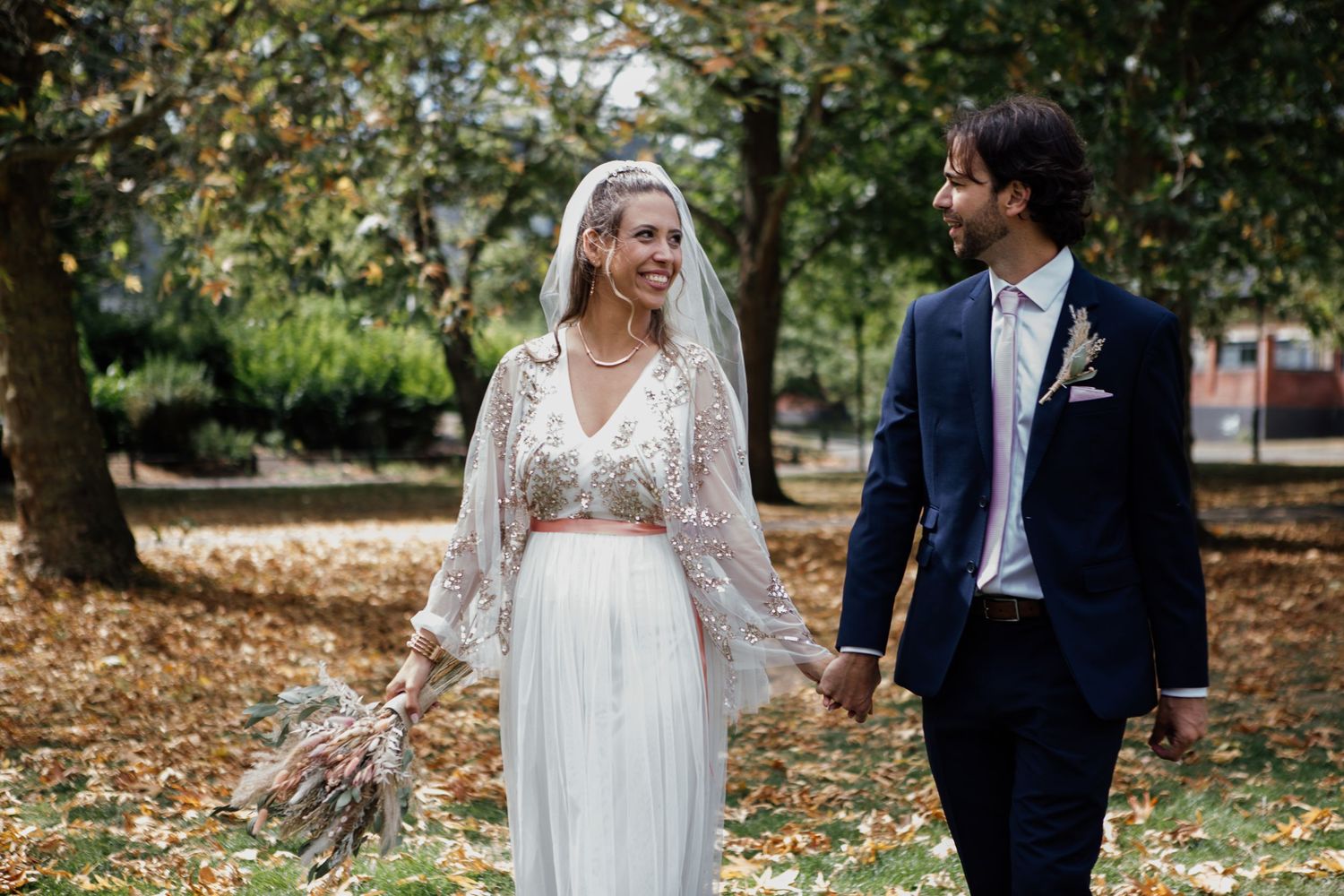Bride and groom walk hand-in-hand through autumn park, bride wearing lace crop top and flowing skirt with veil.