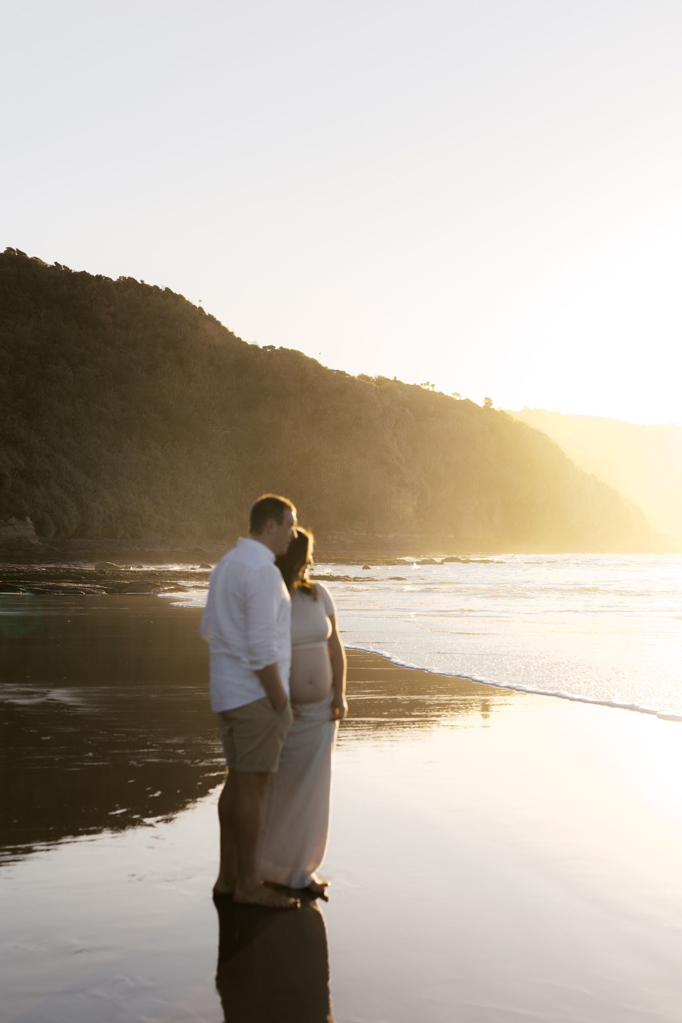 A couple stands together watching the sunset over a scenic beach cove with golden light reflecting on the water.