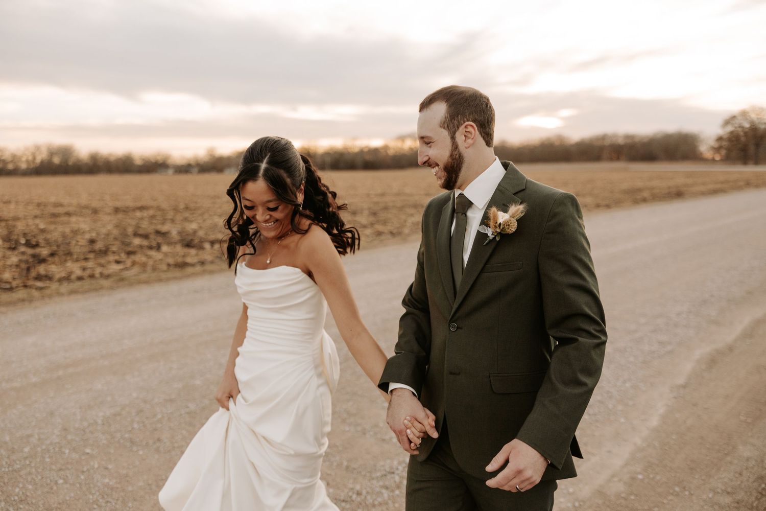 A couple in wedding attire walks hand in hand on a dirt road during sunset in a rural setting.