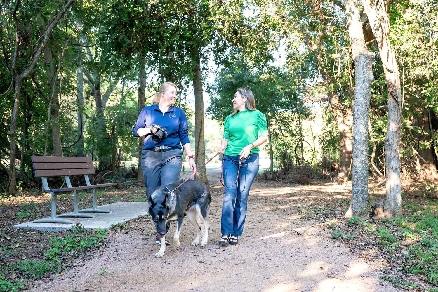 Photographer Kelly J. Russo walks with a client and her dog on a tree-lined trail in a park with a bench visible nearby.