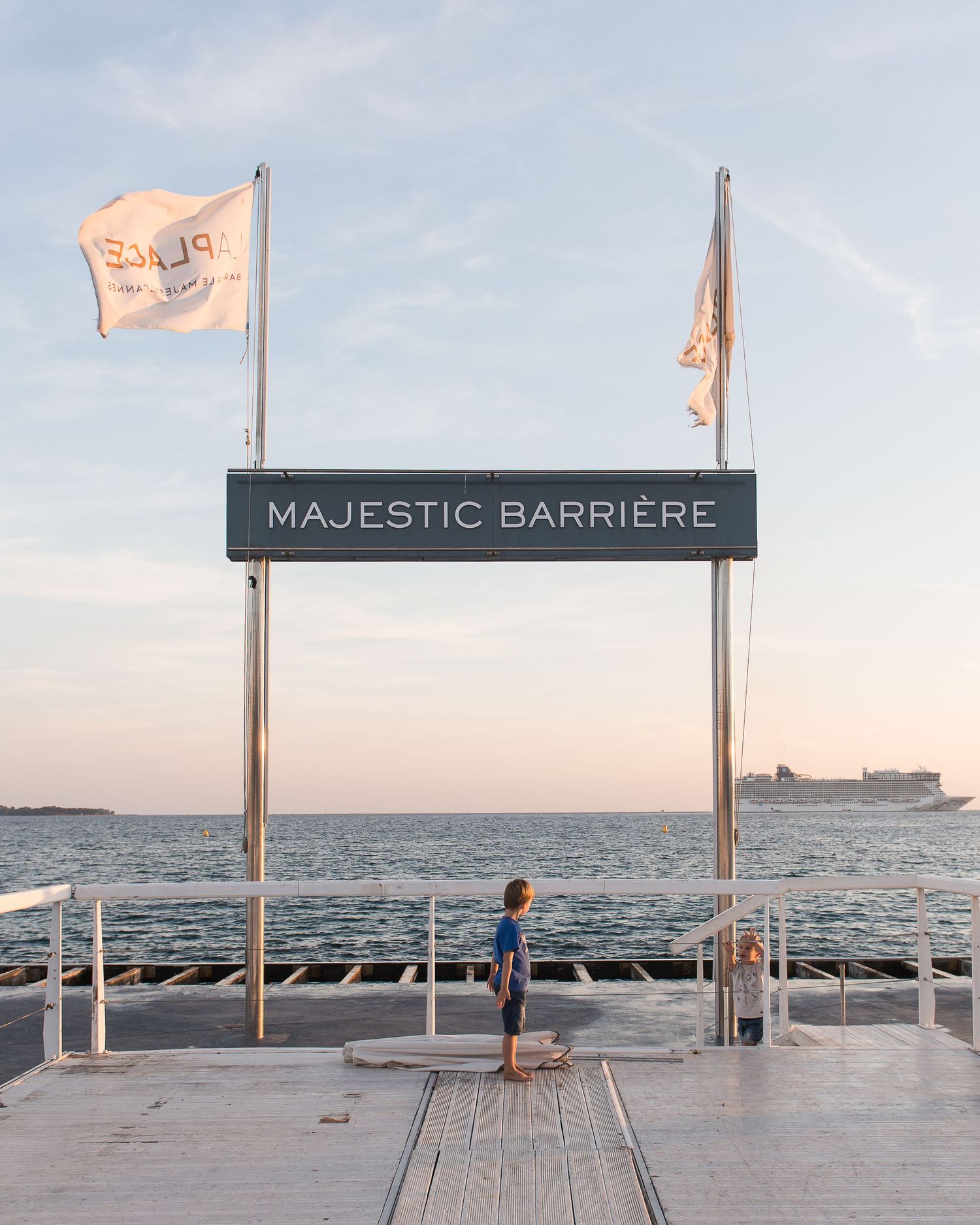 Wooden boardwalk in Cannes with Majestic Barriere sign and flags overlooking the ocean at sunset.