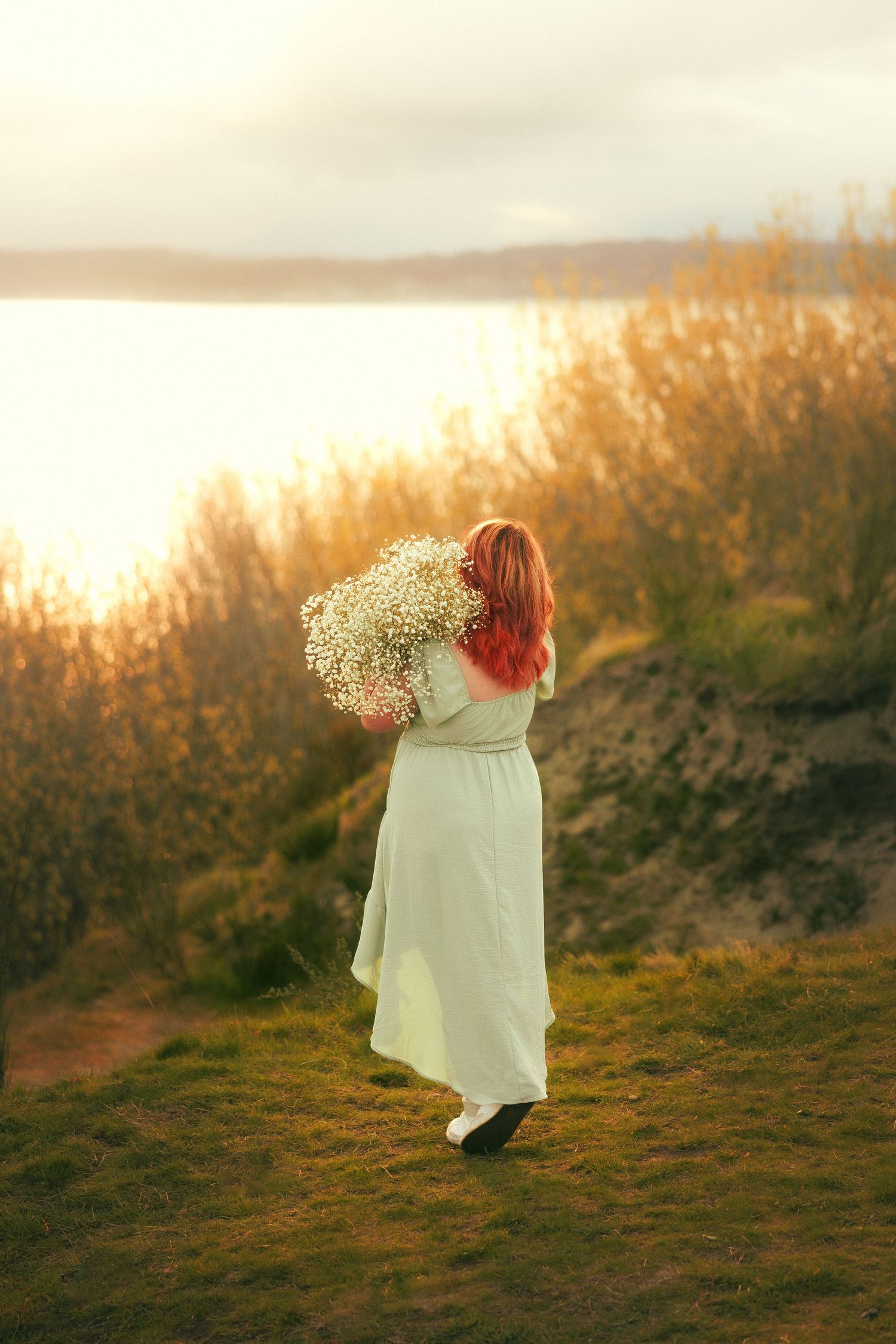 A person in a white dress stands in a golden field at sunset holding a bouquet of baby's breath flowers.
