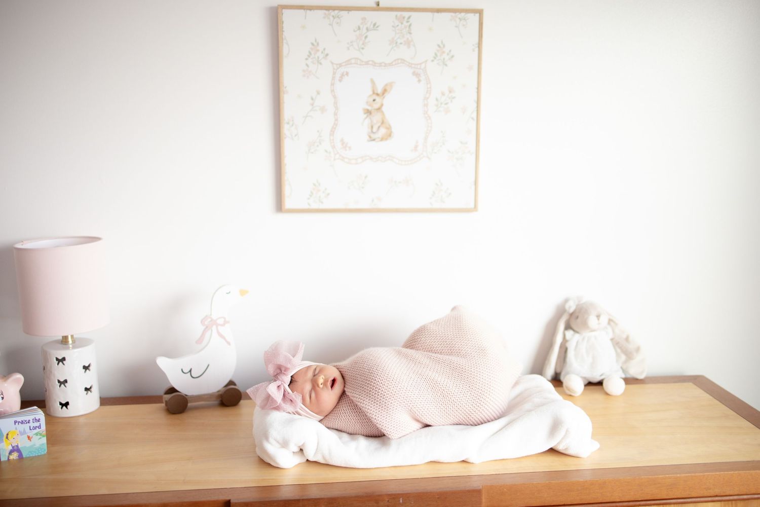 Newborn baby swaddled in pink sleeping peacefully on dresser in bunny-themed nursery with soft decor.