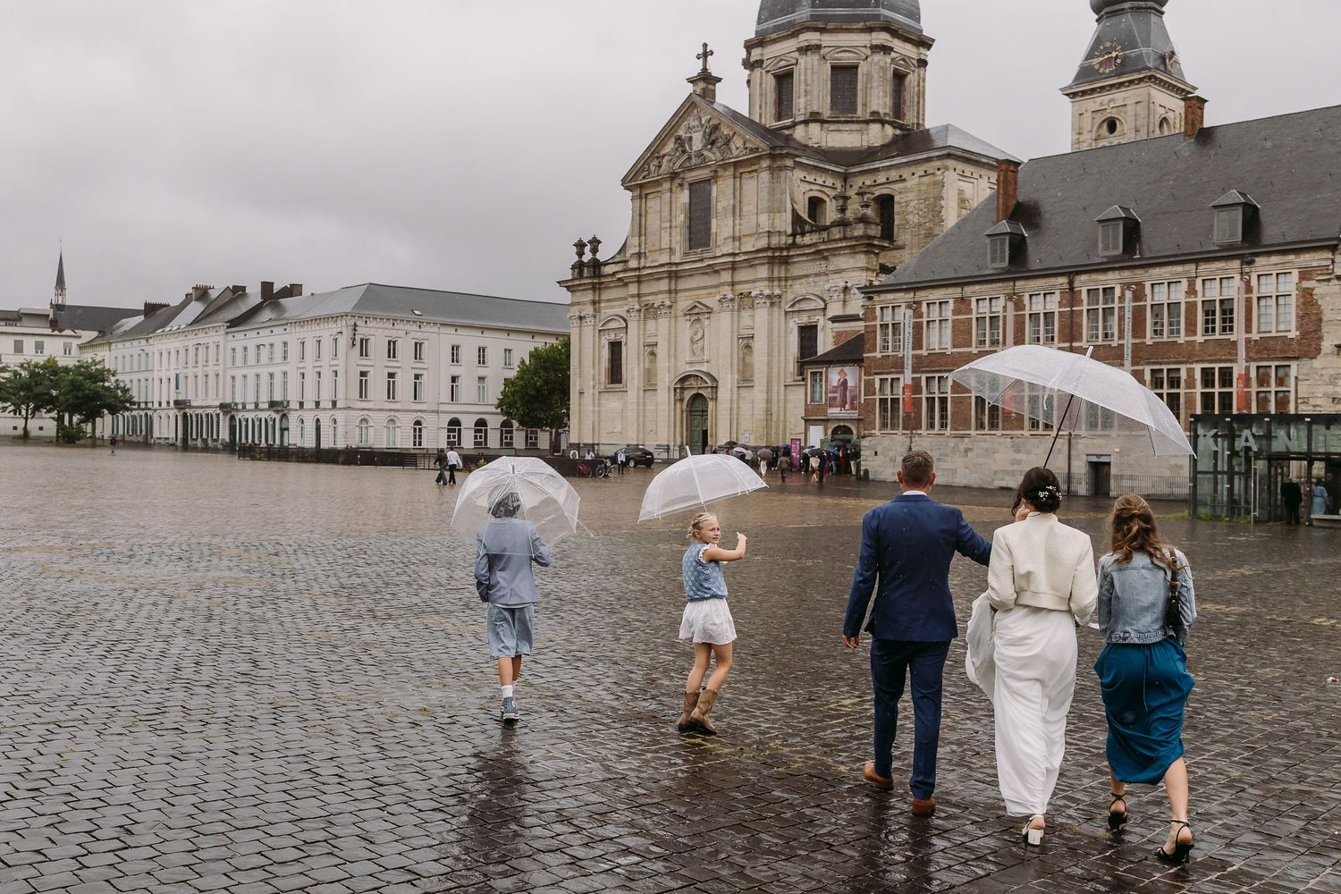Group walks down historic cobblestone street in front of majestic cathedral during rain shower in European city.