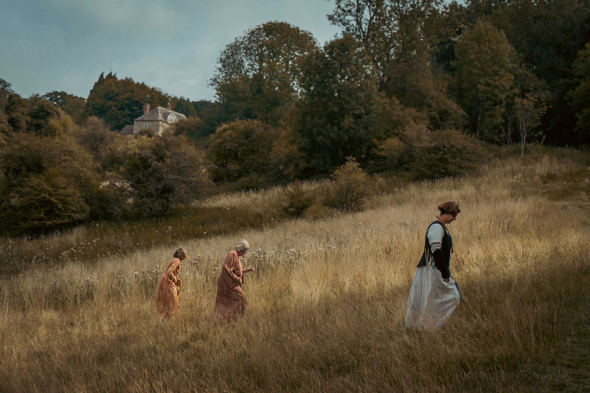 Three figures in period dress walking through a golden autumn field at sunset. Author novel promotional photoshoot in Gloucestershire.
