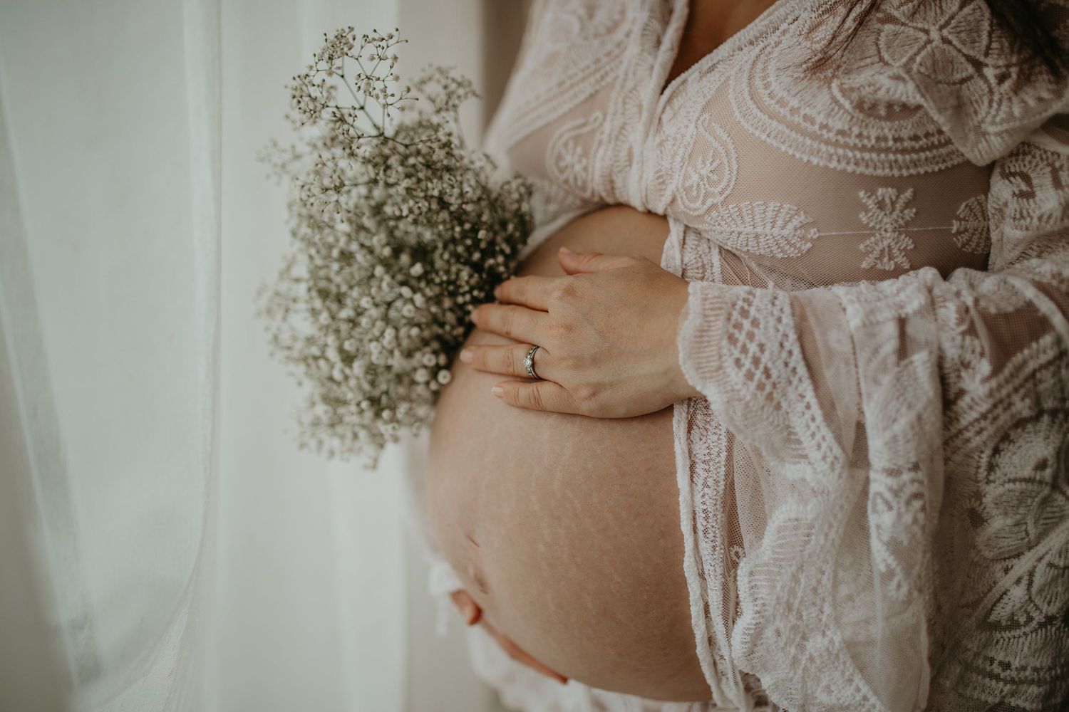 A pregnant belly in white lace dress is gently cradled while holding a delicate bouquet of baby's breath flowers.