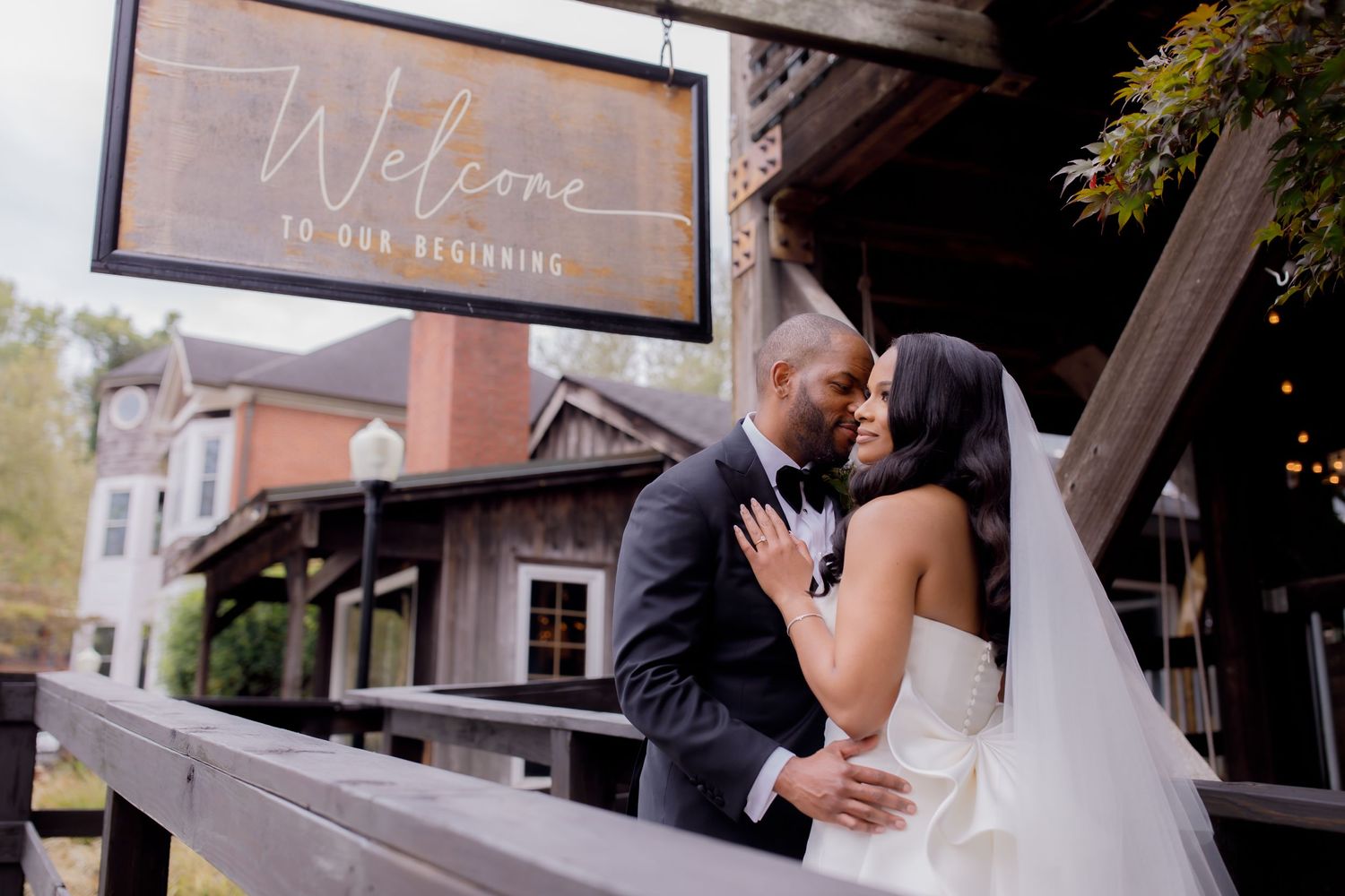 Romantic wedding moment with couple embracing under a welcome sign at rustic venue entrance.
