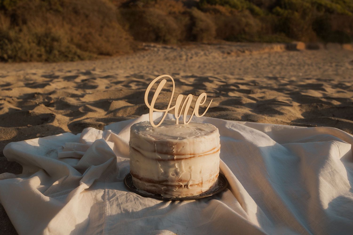 Rustic beach setting with small cake topped with 'One' wooden topper on white fabric.