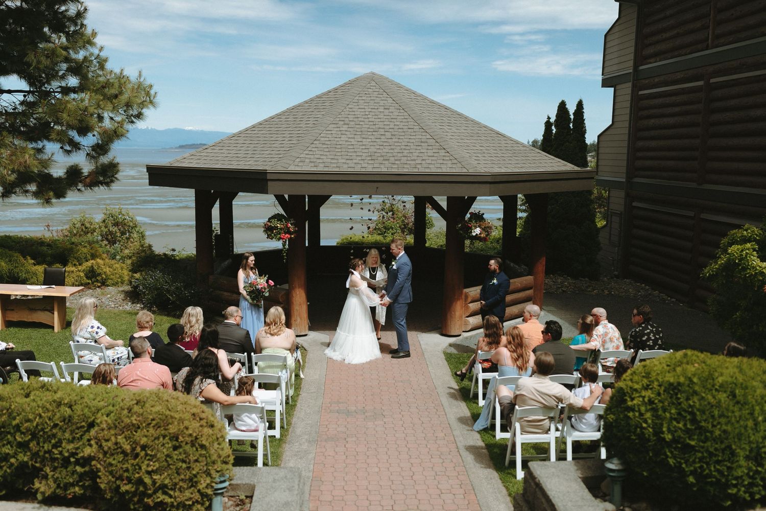 An outdoor wedding ceremony takes place under a covered gazebo overlooking the water with guests seated on white chairs.