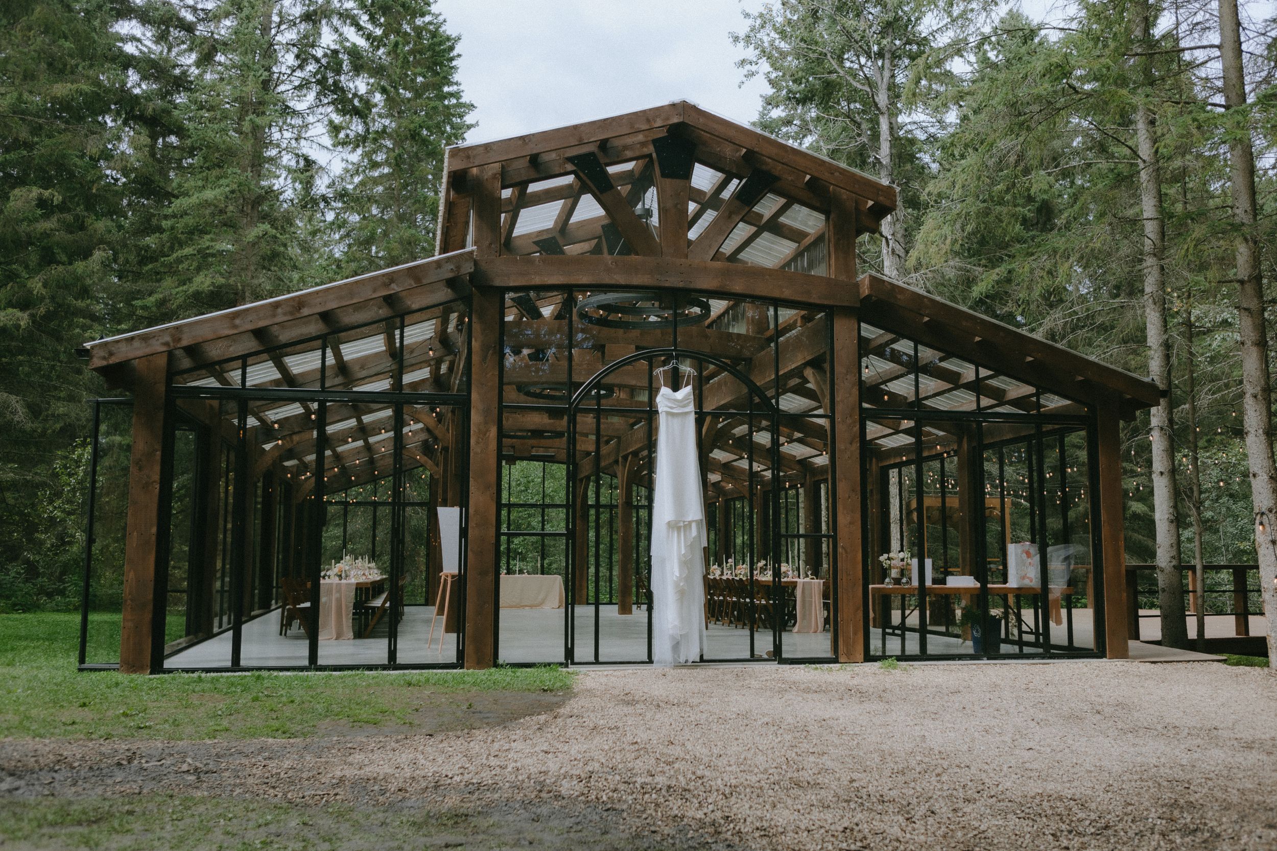 A modern glass and wood wedding pavilion surrounded by evergreen forest.