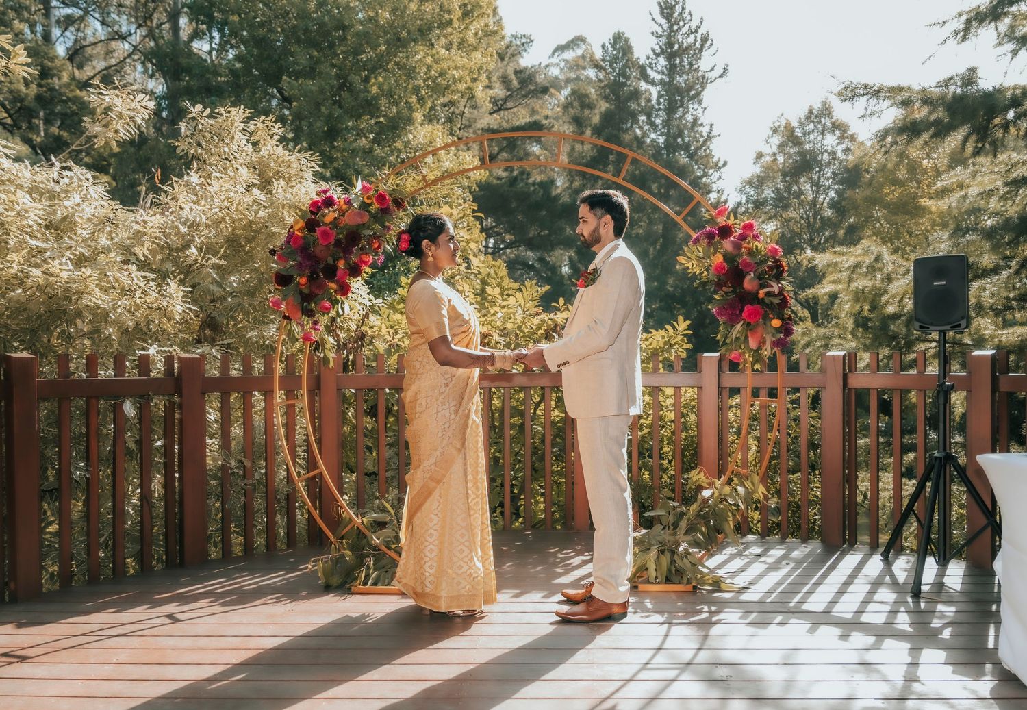 Two people holding hands walking through a field at sunset wearing formal attire with person holding floral bouquet.