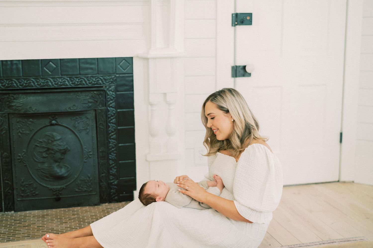 A mother in a white dress sits on a wood floor near an ornate dark green fireplace, holding and gazing at her baby.