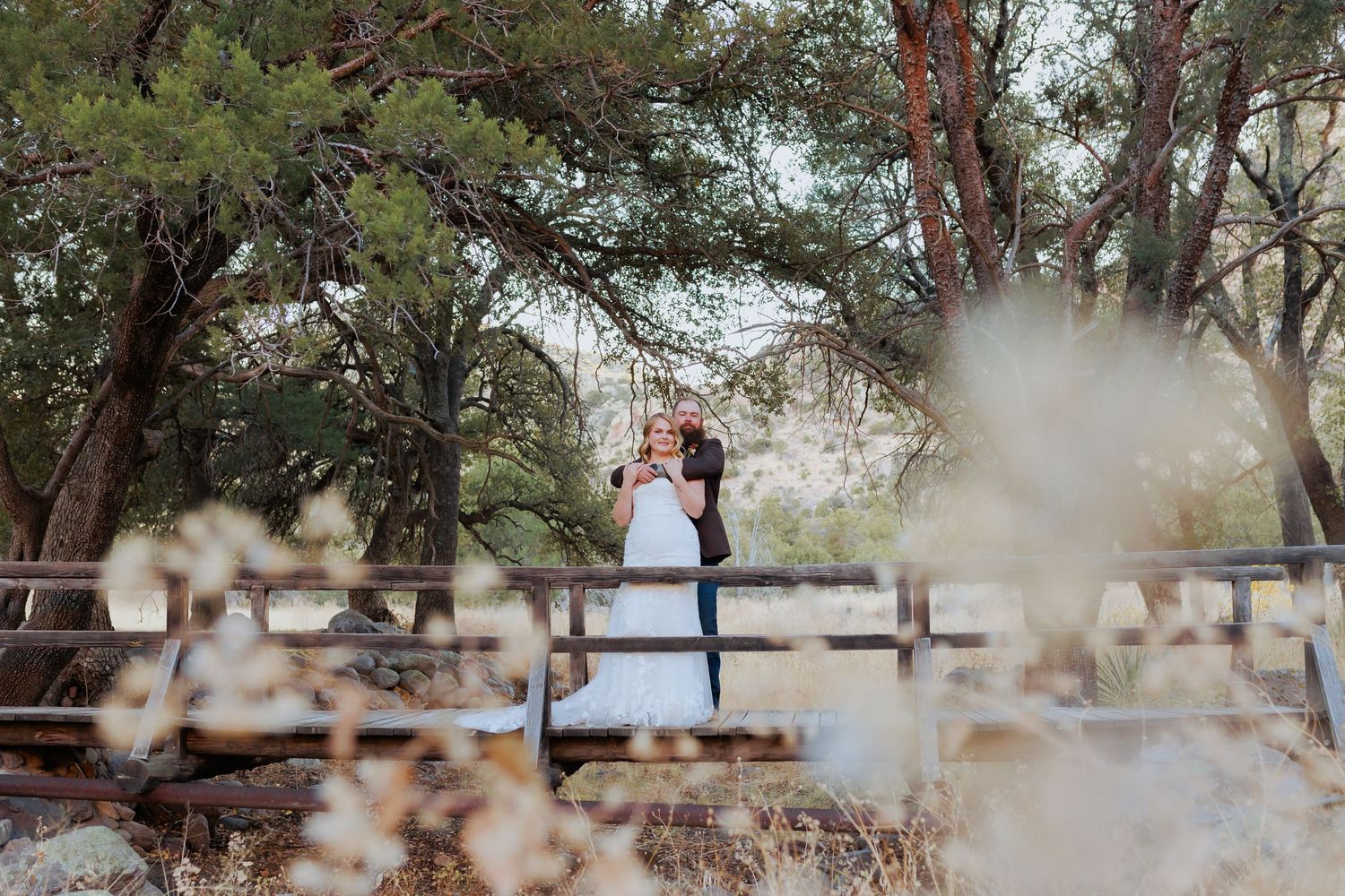 A couple embraces on a wooden bridge surrounded by pine trees and soft bokeh at dusk. Wedding photography Willcox, Arizona.