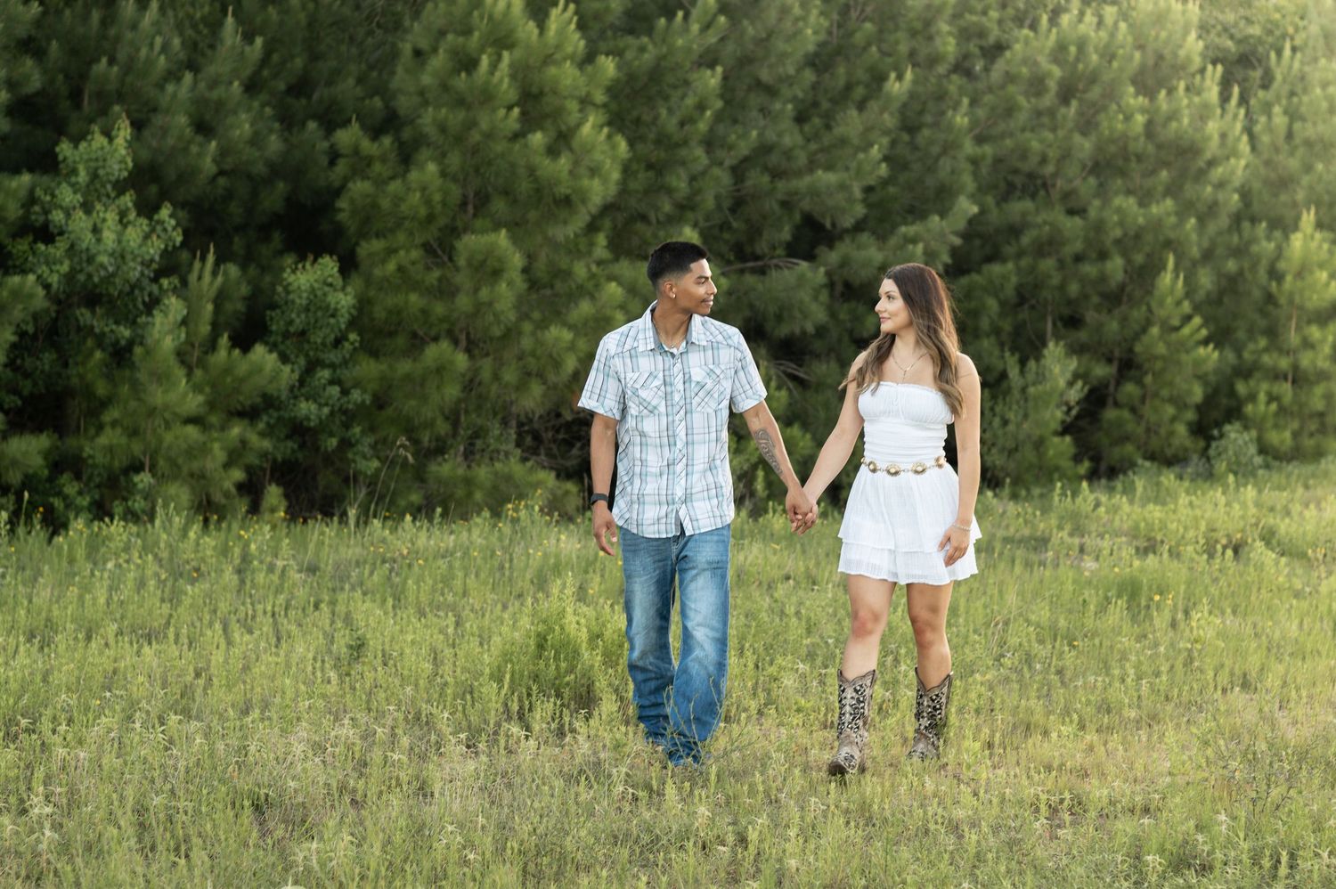 A couple holds hands while walking through a grassy meadow with pine trees in the background.