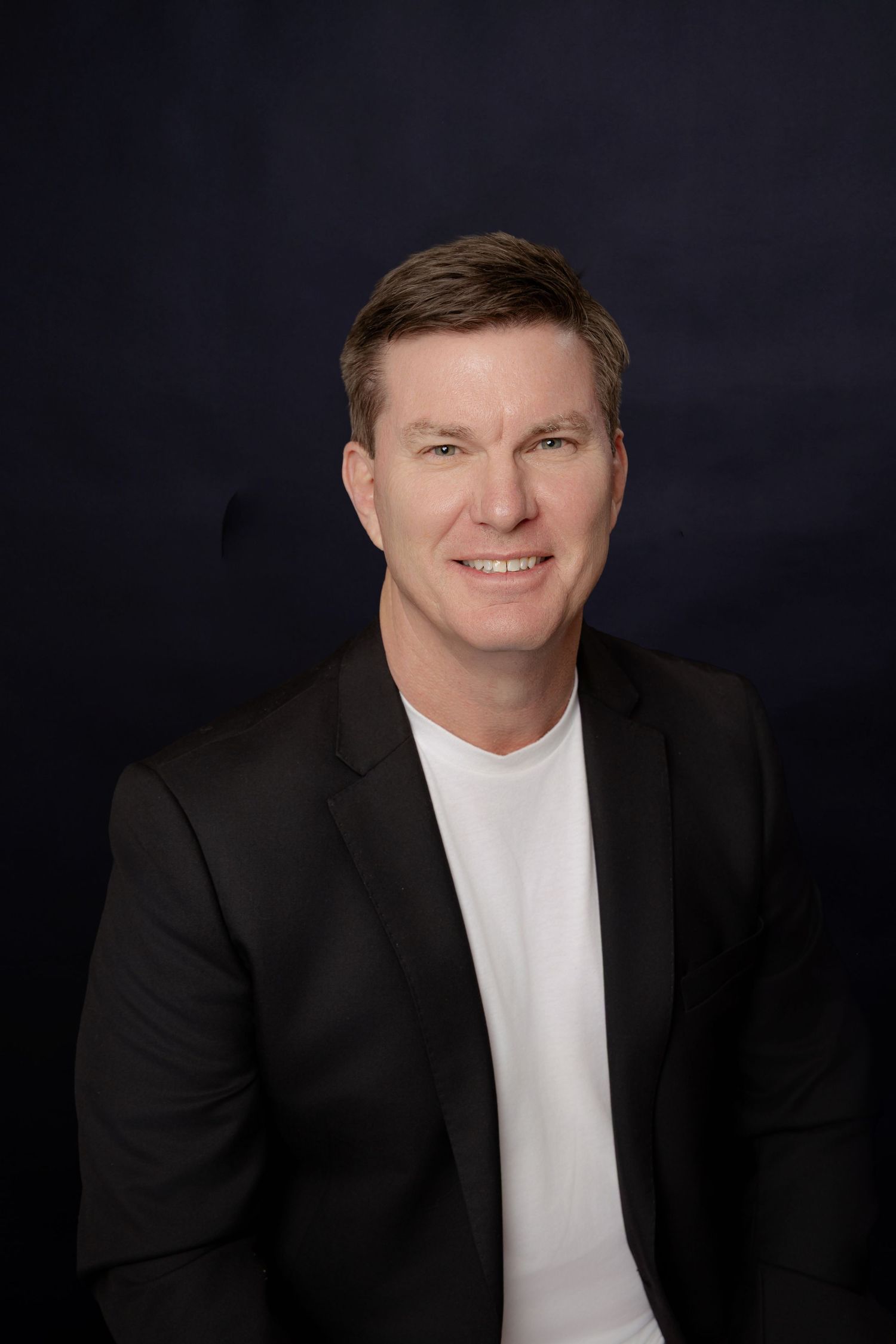 Professional headshot of person in black blazer and white t-shirt against dark background.
