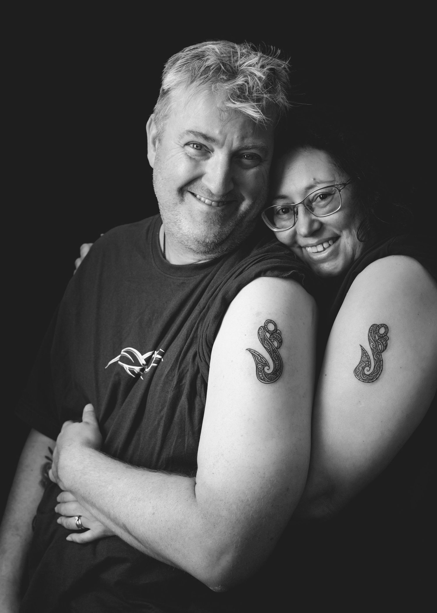 A black and white portrait of two smiling people showing off their matching seahorse tattoos on their arms.