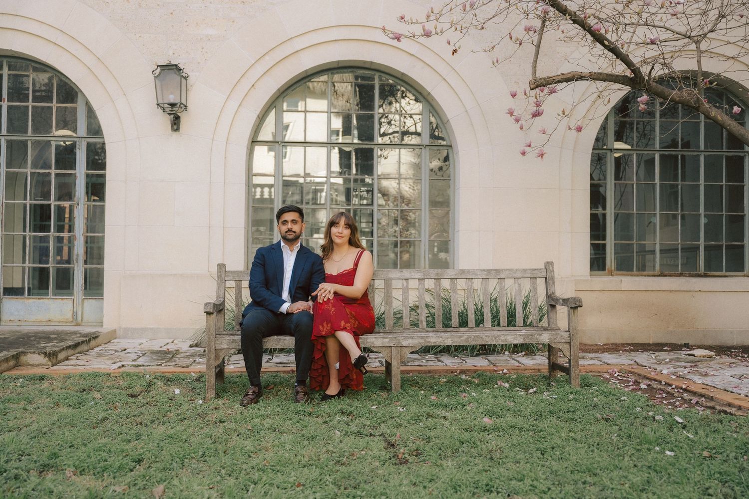 A couple sits together on a wooden bench in front of an elegant arched window with a white stone facade.