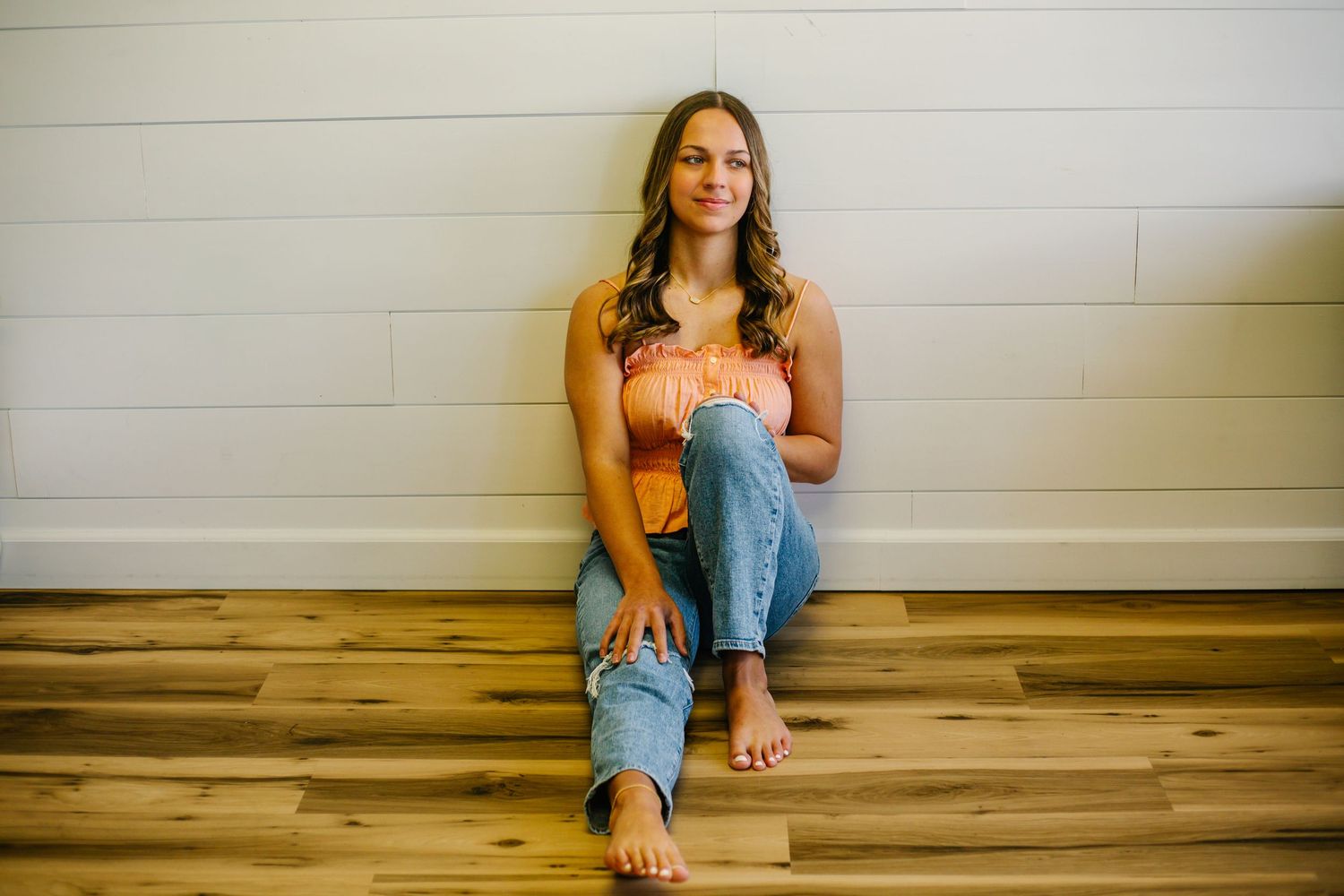 Person sitting on hardwood floor wearing ripped jeans and orange shirt against white wall.