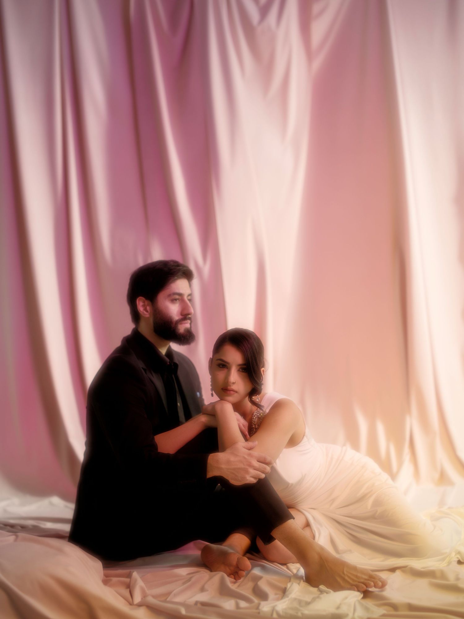 Romantic couple poses intimately against soft pink draped fabric backdrop in dreamy portrait session.