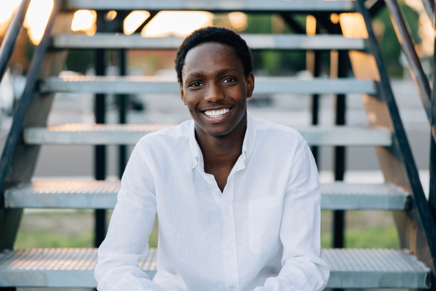 Smiling young Black man in white shirt sitting on metal stairs outdoors at sunset.