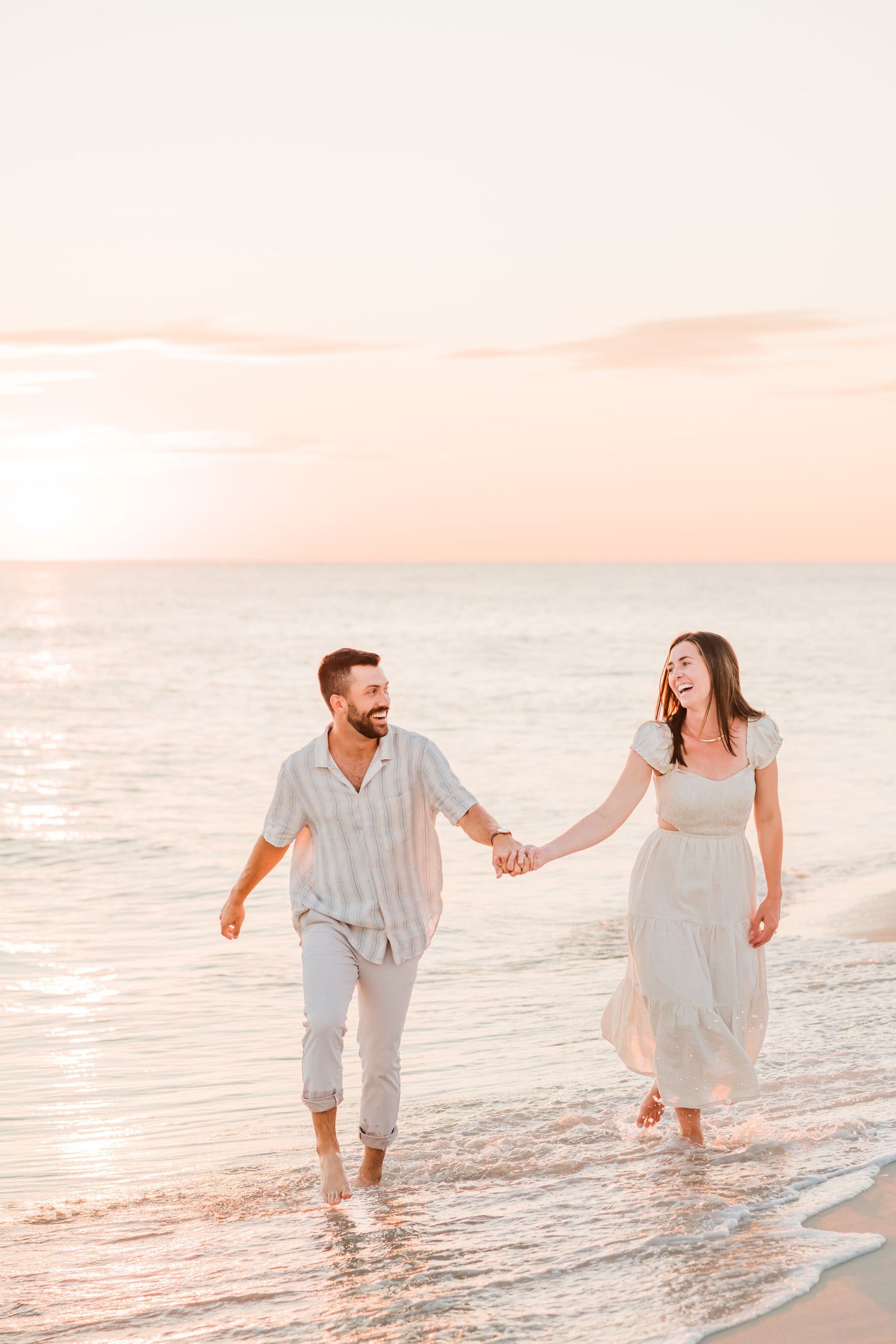 A couple in light-colored outfits walks hand in hand along a beach at sunset, laughing together by the ocean waves.