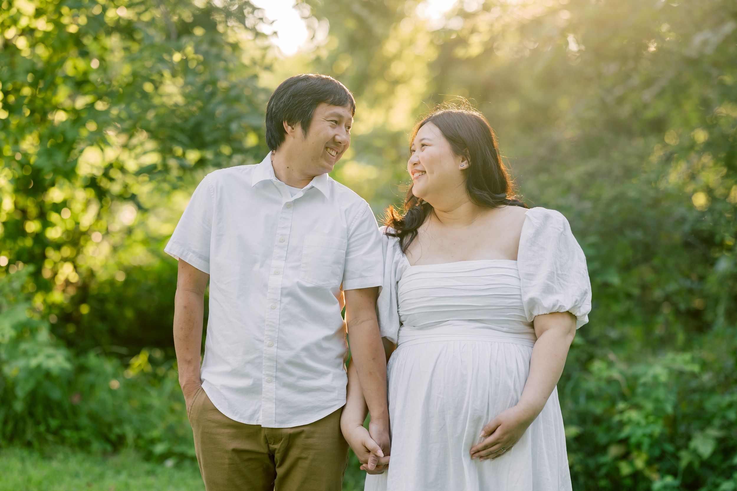 Happy couple holding hands outdoors in a sun-dappled garden setting, wearing white casual clothing.