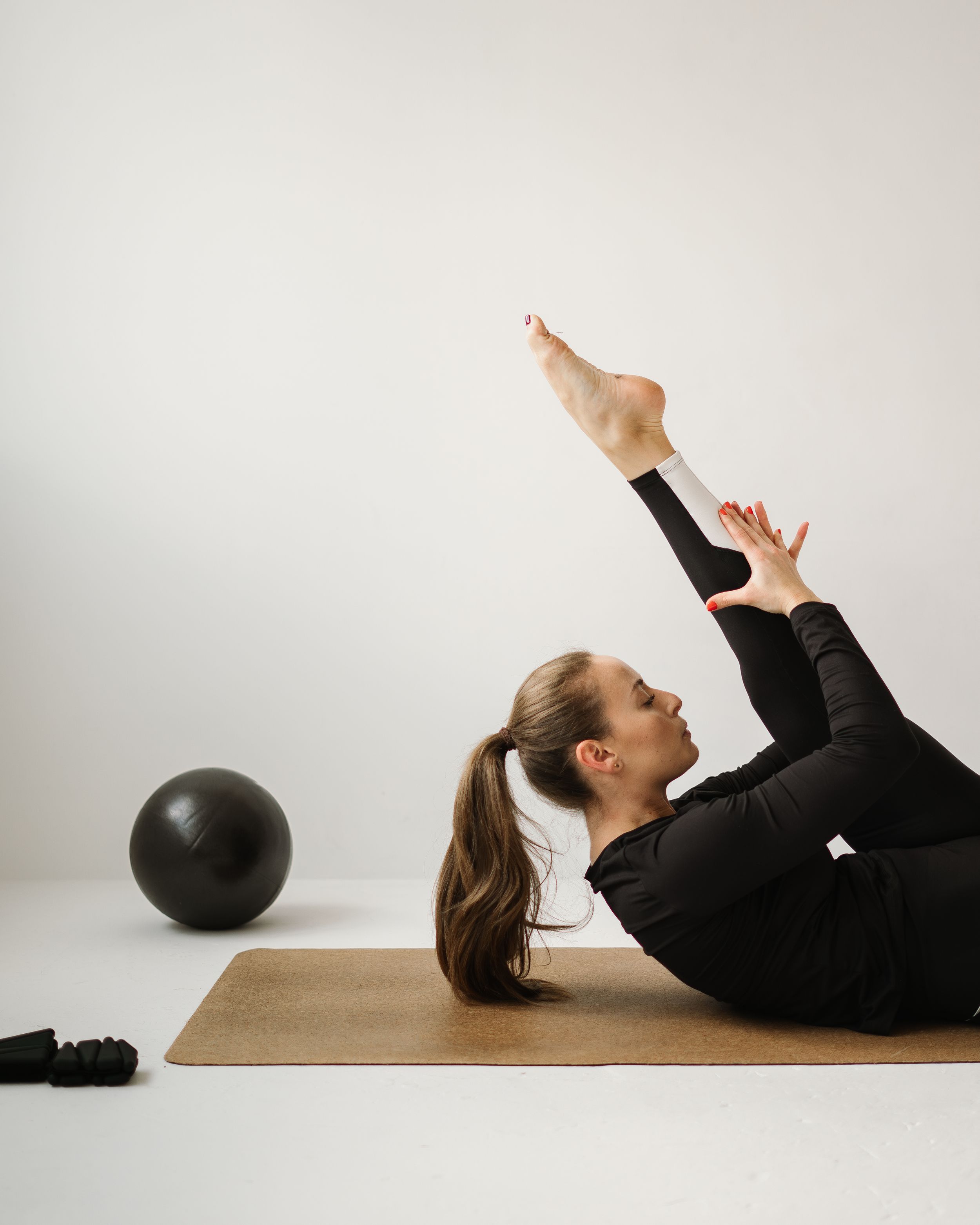 Woman in black athletic wear practicing pilates core exercises on a mat with a fitness ball nearby on white background.