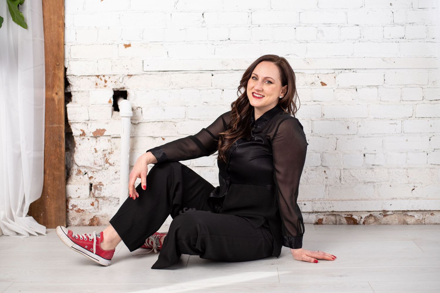 Casual figure sits relaxed on floor wearing black outfit and red sneakers against white brick wall backdrop.