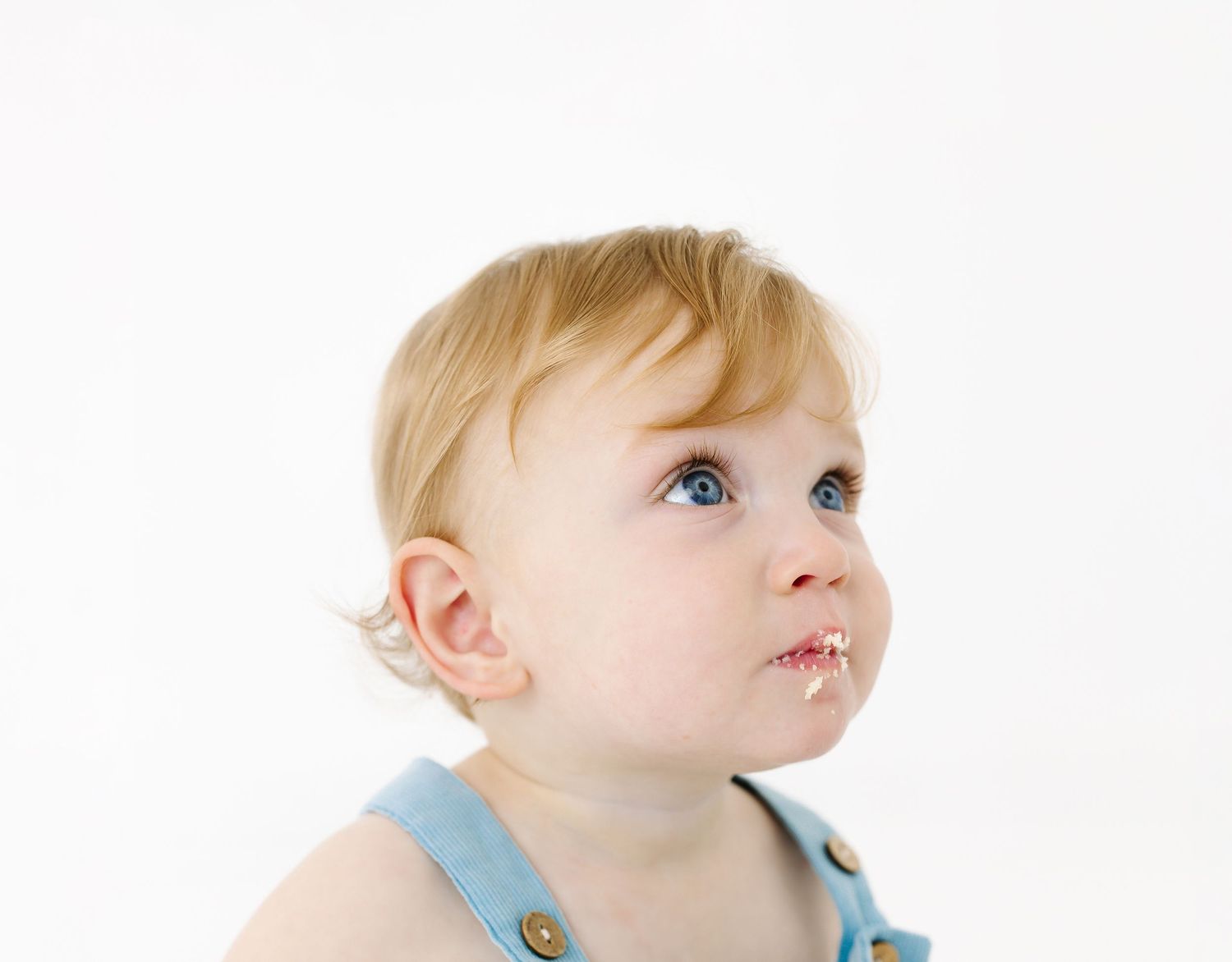 Young child in blue overalls gazes upward with bright blue eyes against white background.