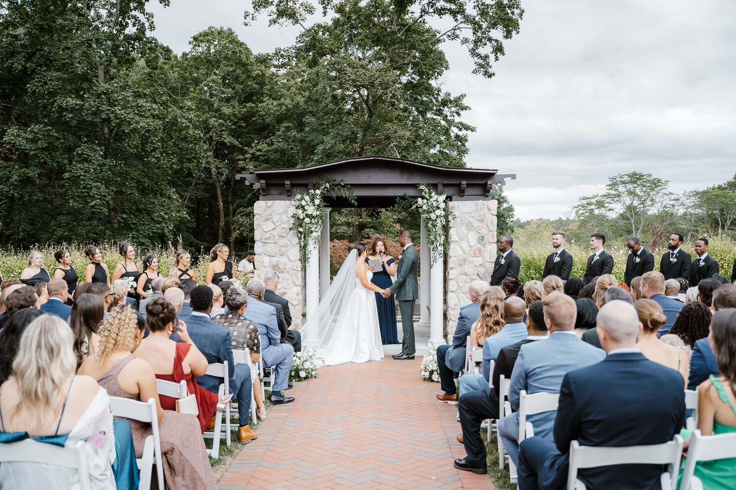 Outdoor wedding ceremony under a decorated arch with guests seated on white chairs.