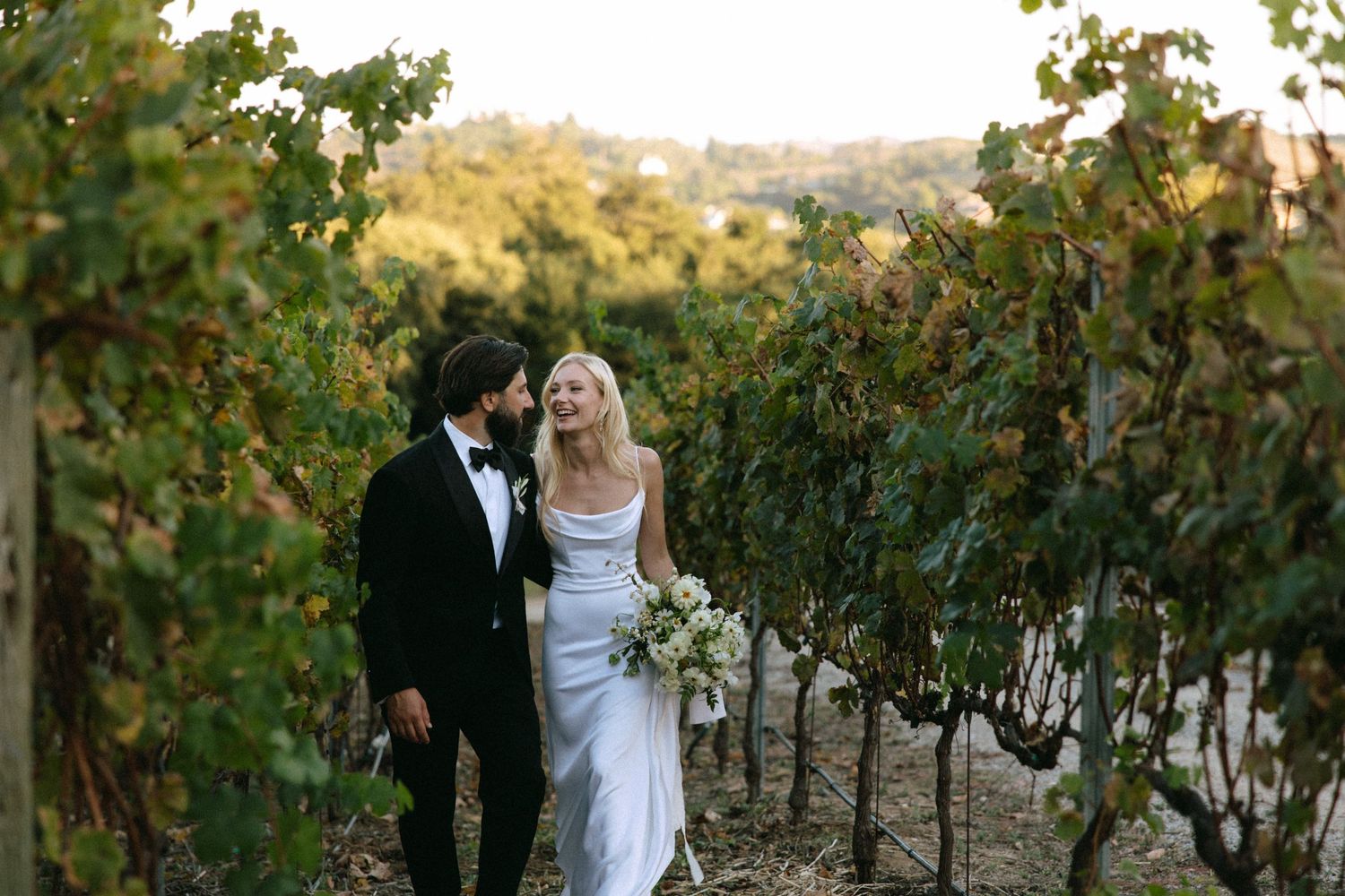 Bride and groom walking through vineyard rows in wedding attire with bridal bouquet.