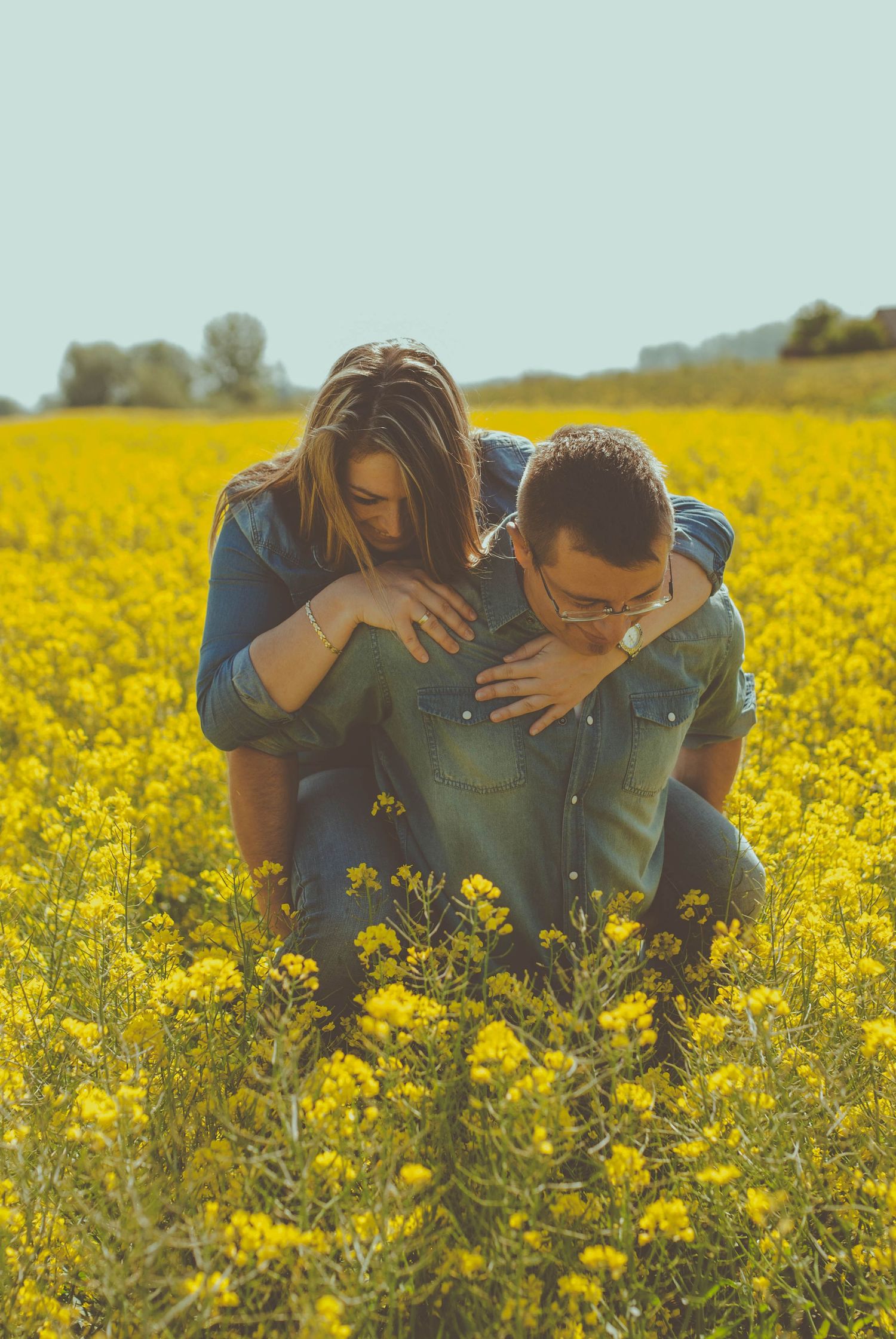 Two figures embracing in a vibrant yellow canola field on a sunny day with rolling hills in the background.