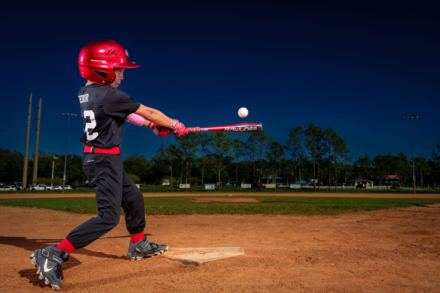 Youth baseball player in red helmet swings bat to hit ball on dirt infield at dusk with trees in background.