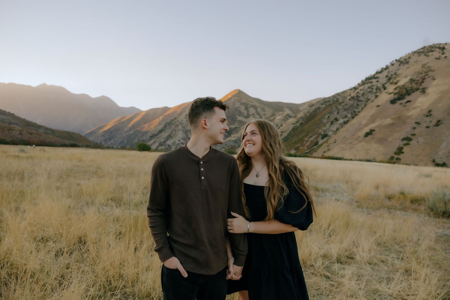 Couple walking through golden grass fields with stunning mountain backdrop at sunset.