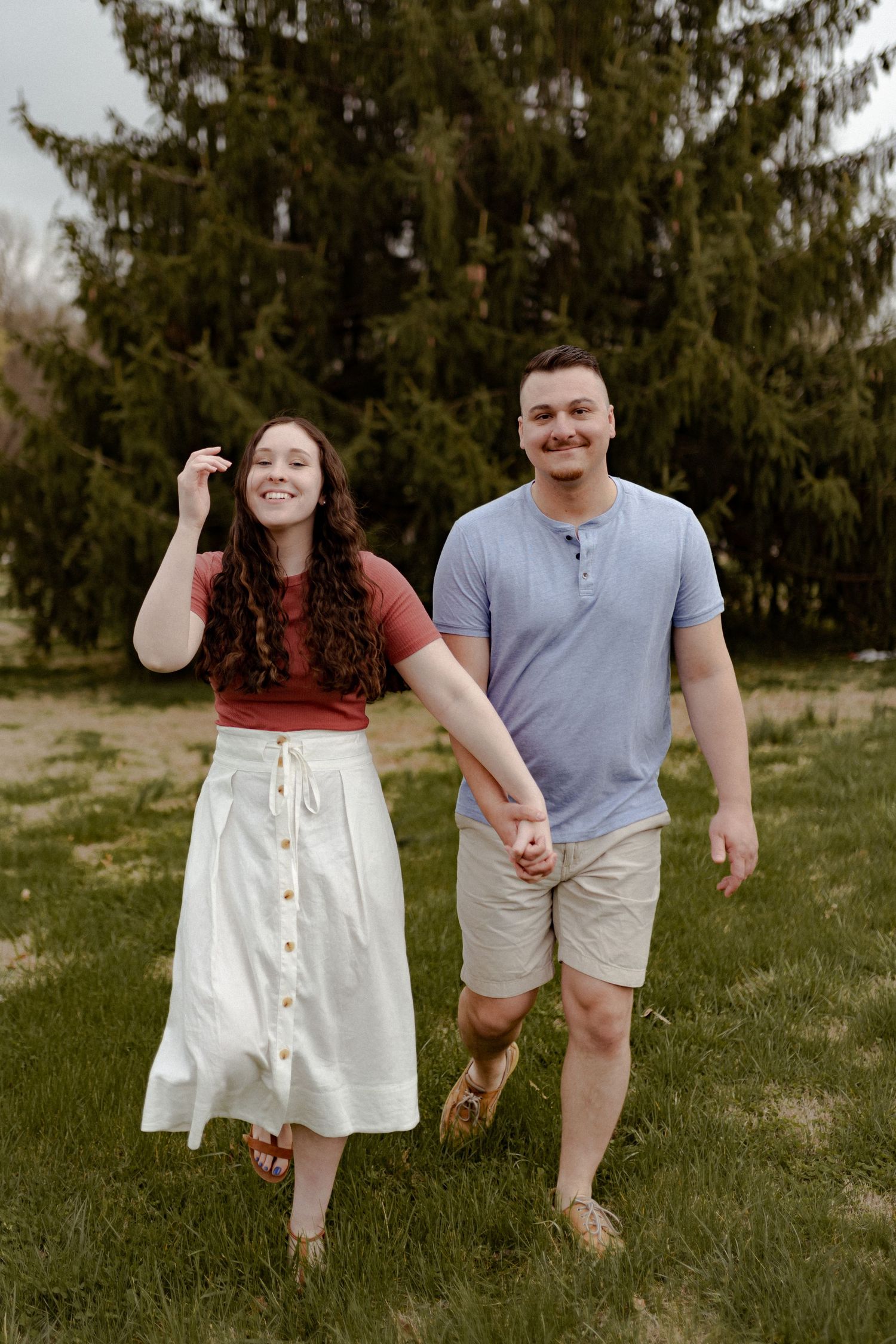 Casual couple walking hand in hand through grassy field with evergreen trees in background.