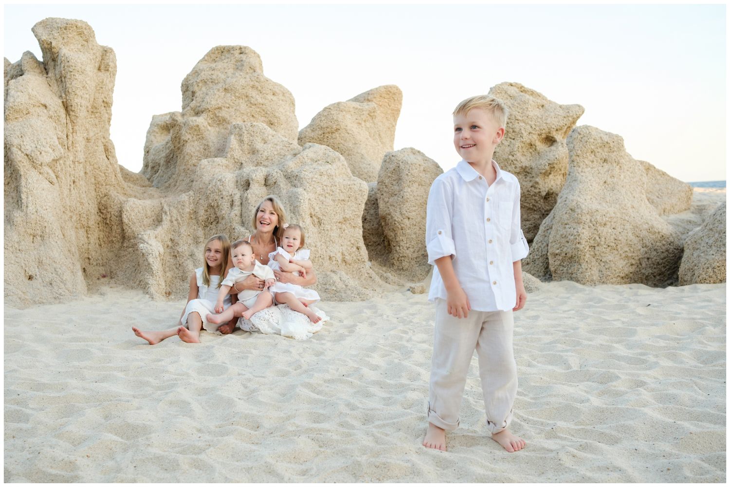 Beachgoers in white and pastel clothing gather near large rocky formations on a sandy beach during golden hour.