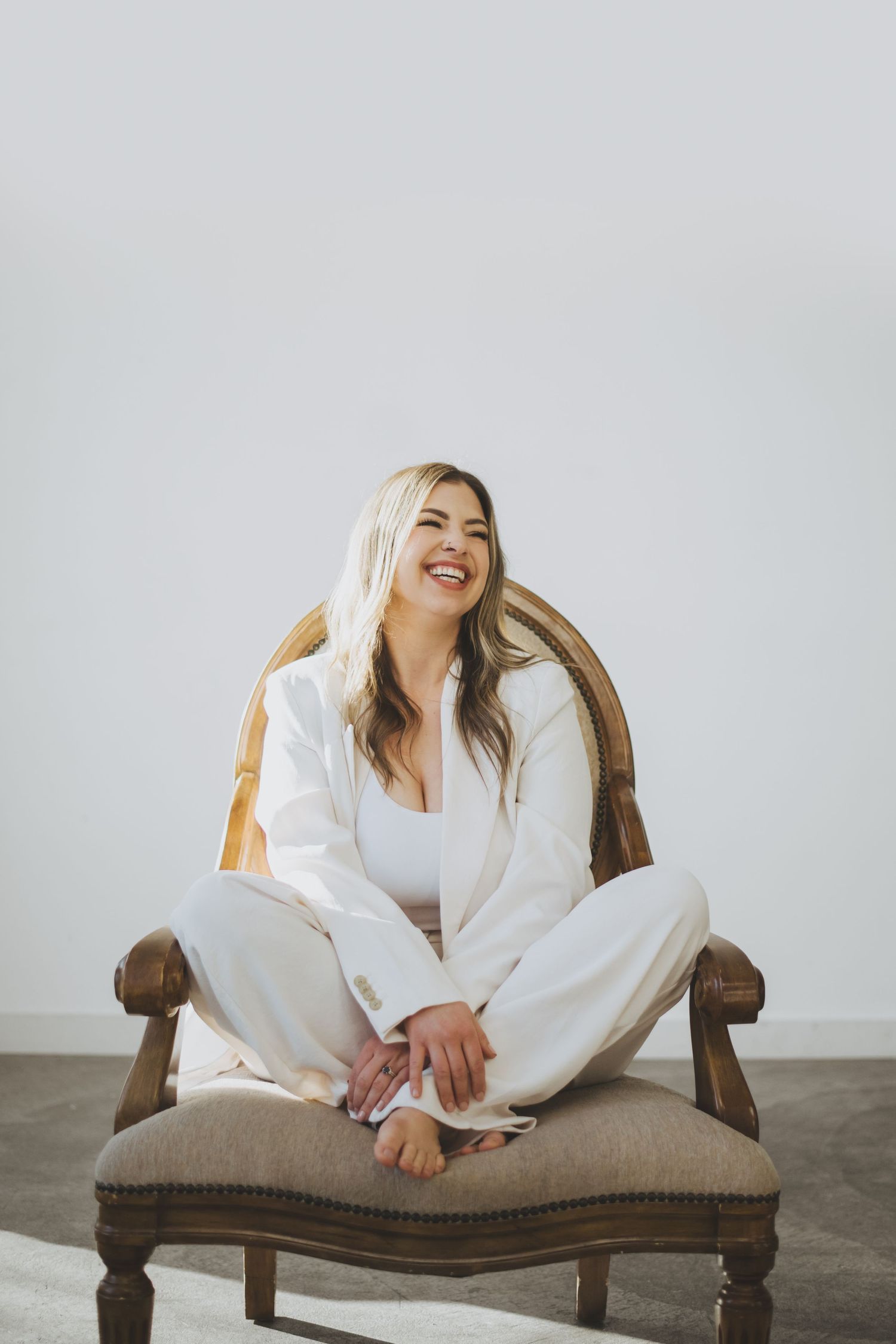 A joyful woman wearing all white lounges comfortably in a vintage brown upholstered chair against a light gray background.