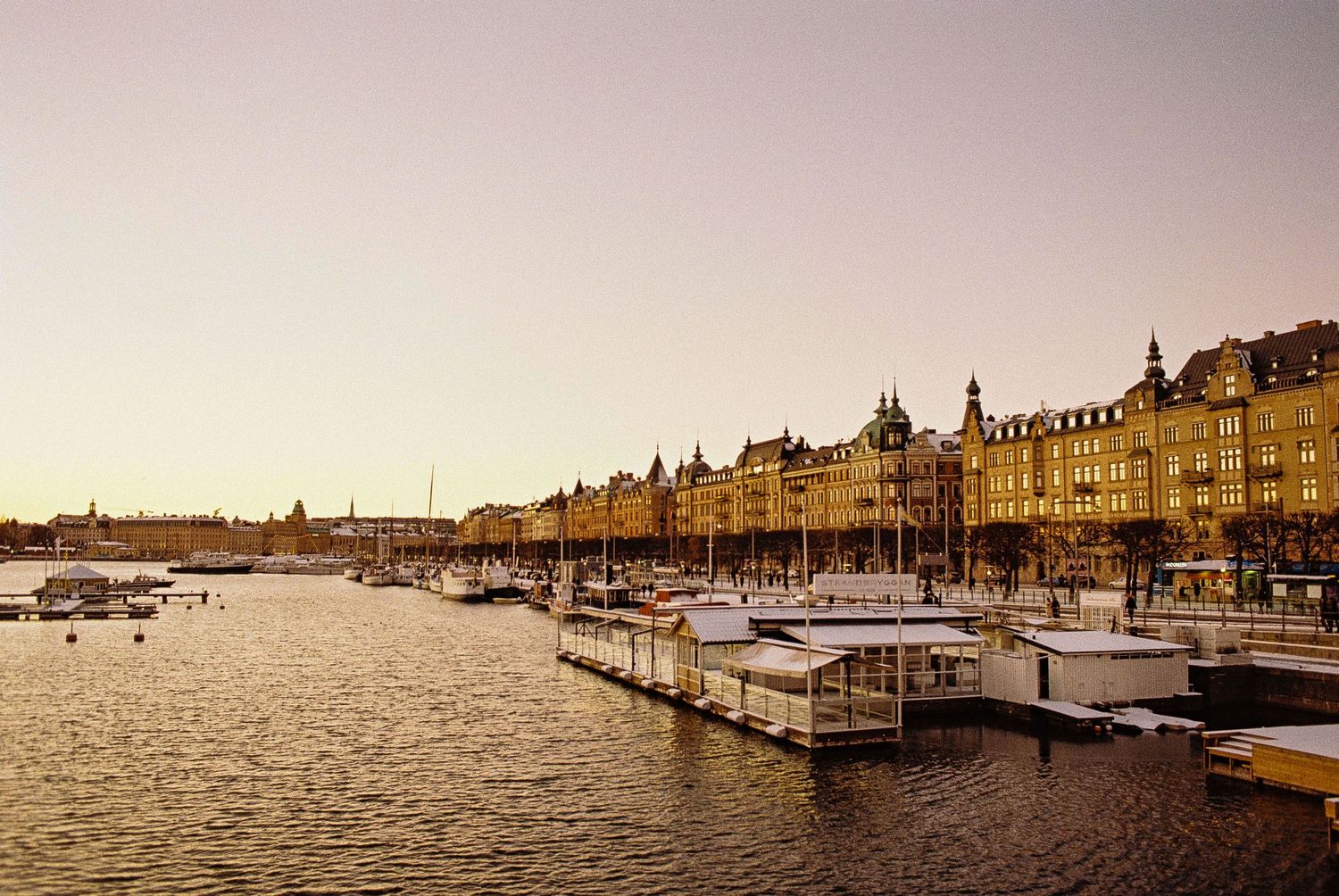 Sunset view of Stockholm waterfront with boats docked along historic buildings and golden light reflecting on calm waters.