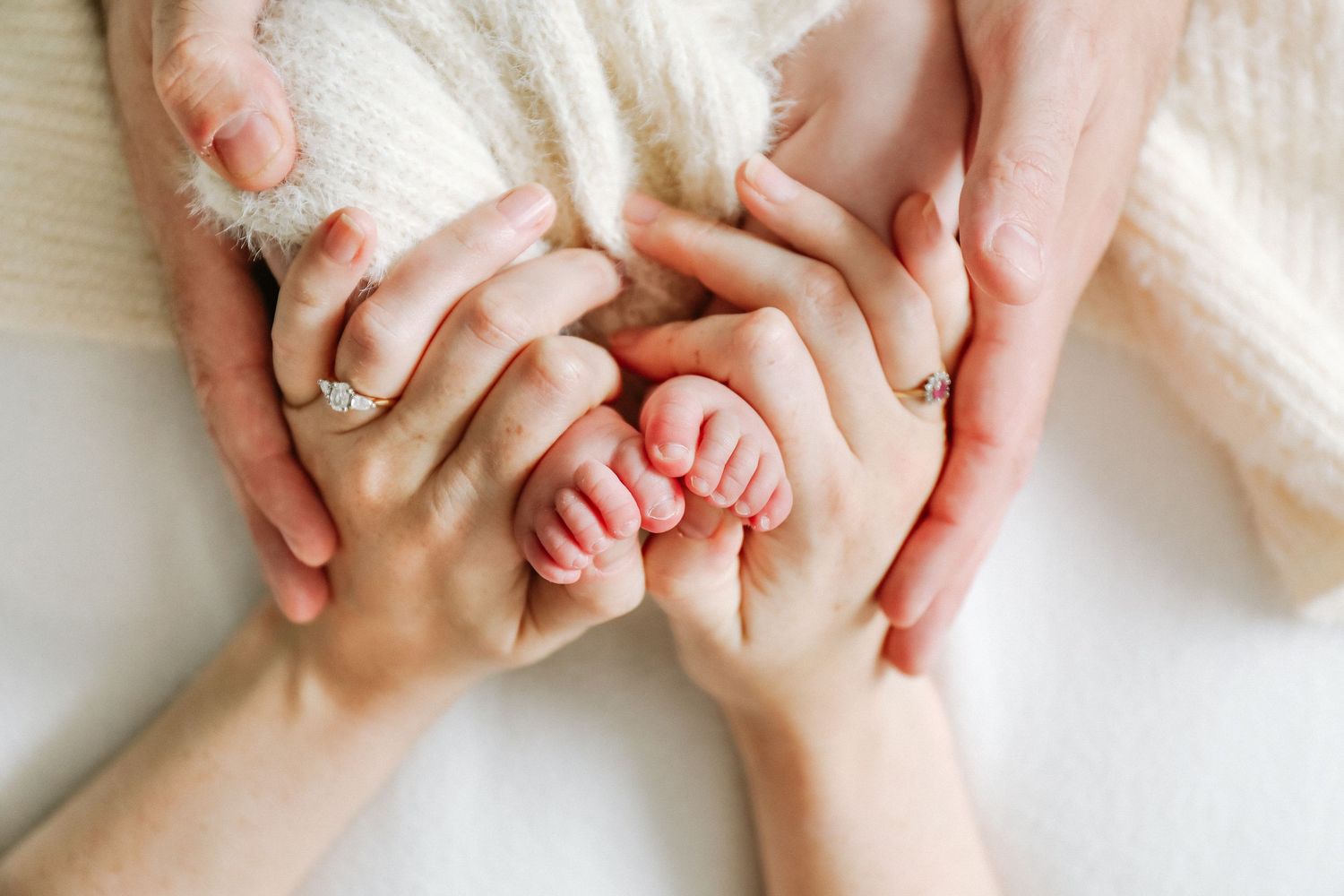 Tiny baby feet nestled between adult hands against a soft white blanket background,during newborn photoshoot in Nottingham