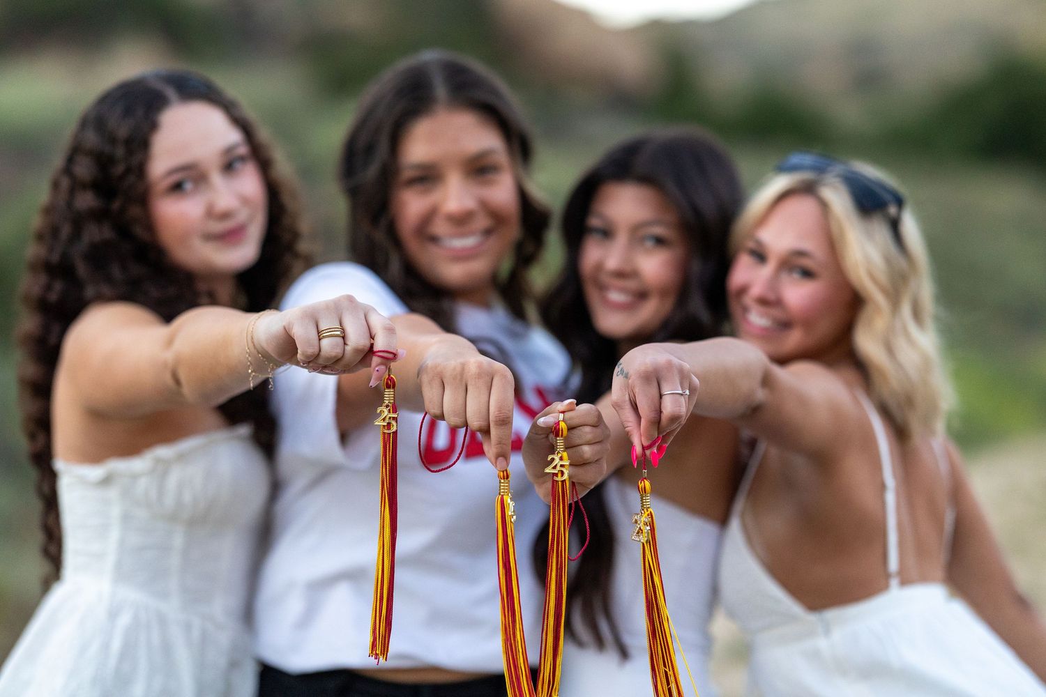 A group in white dresses holds graduation tassels together in front of a blurred outdoor background.