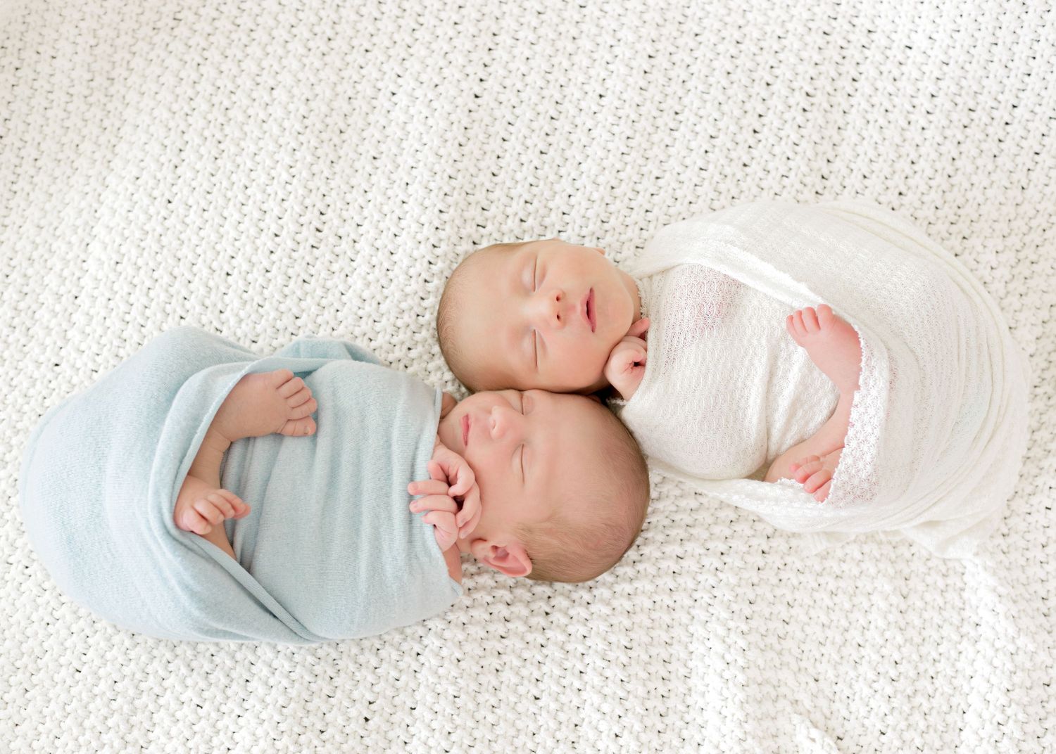 Twin newborn boys wrapped in swaddles while laying on a white blanket. One boy in a blue wrap and the other in a white wrap.