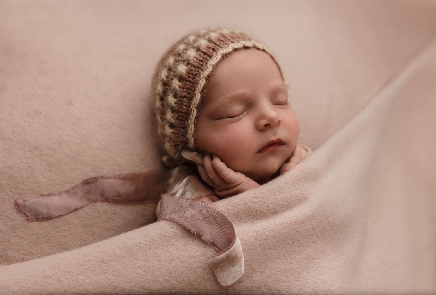 Baby girl in a pink bonnet sleeping peacefully on a blush blanket with her hands resting on her cheek, Charlottesville newborn photographer.