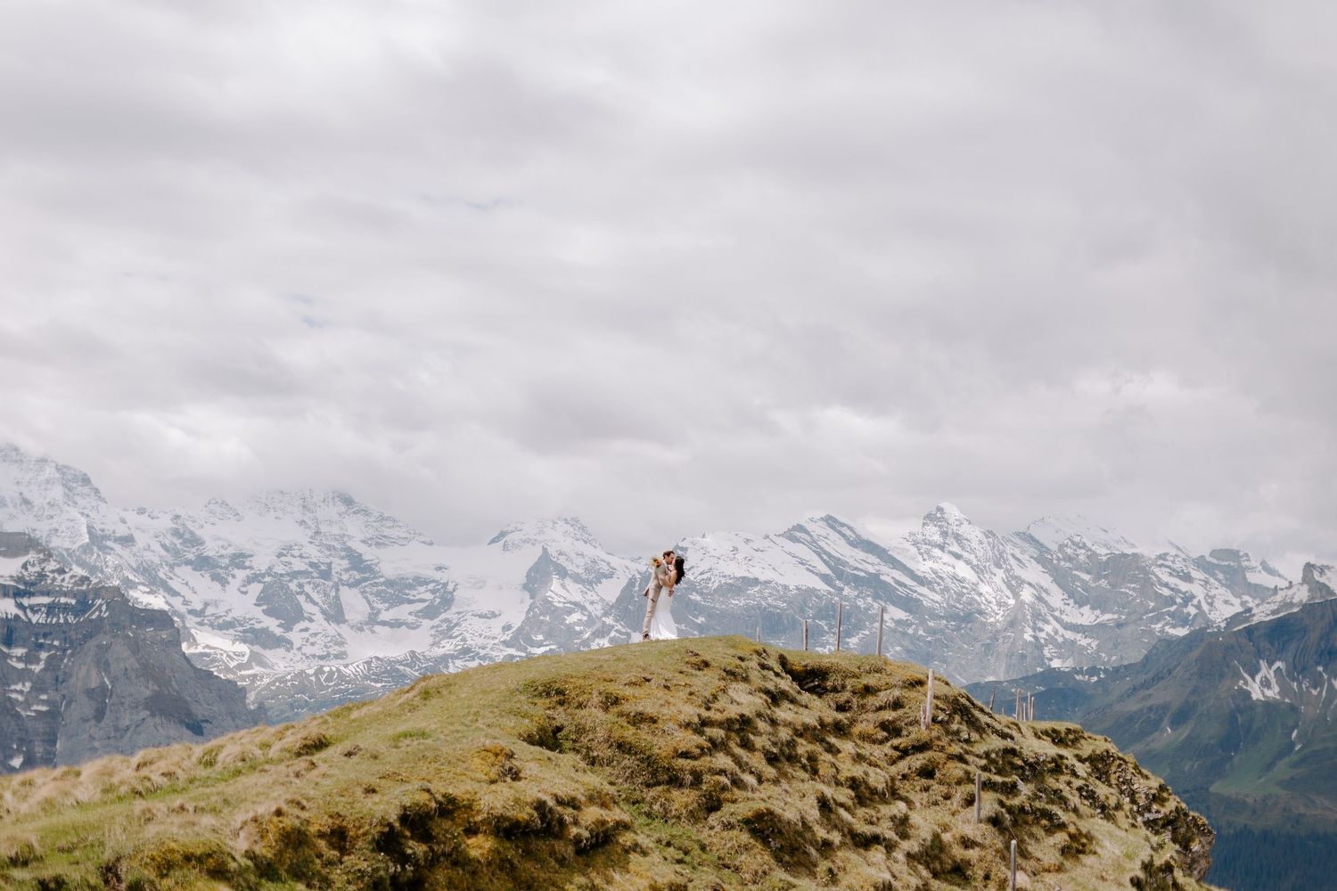 A couple shares a romantic moment atop a grassy mountain peak with dramatic mountain vistas and flowing veil in the breeze.