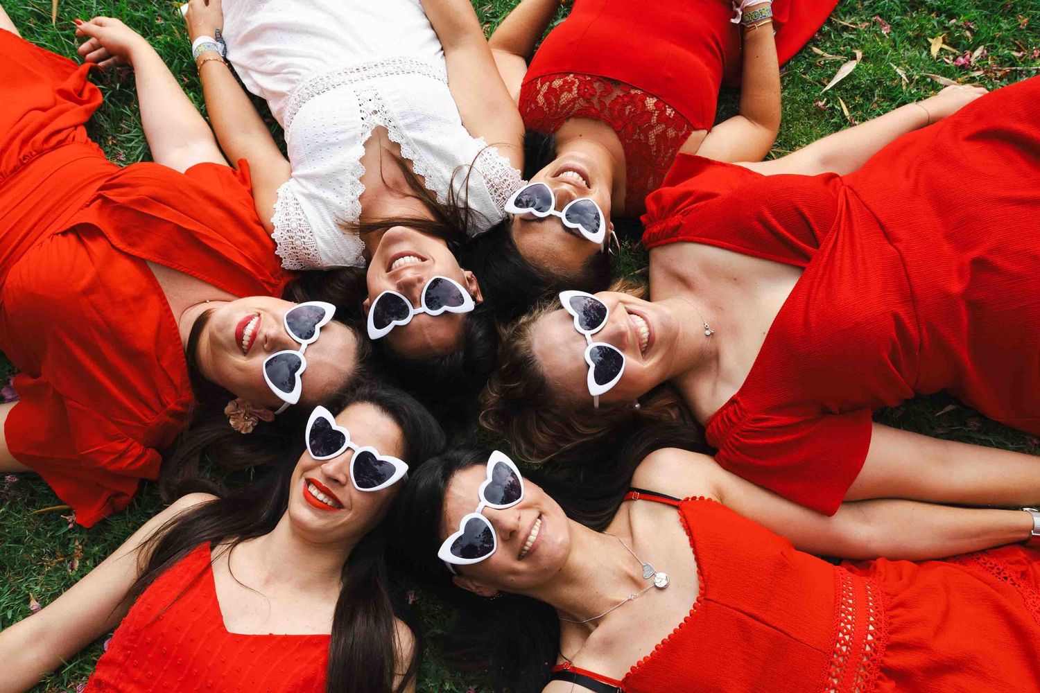Birds eye view of people laying in grass wearing matching heart shaped sunglasses and red dresses.