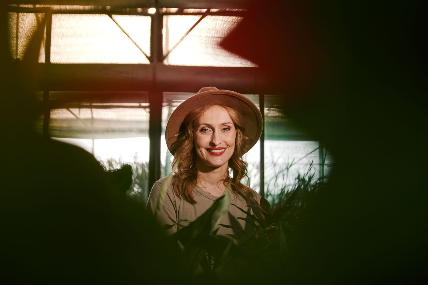 Smiling woman wearing a hat surrounded by lush green plants in warm light.