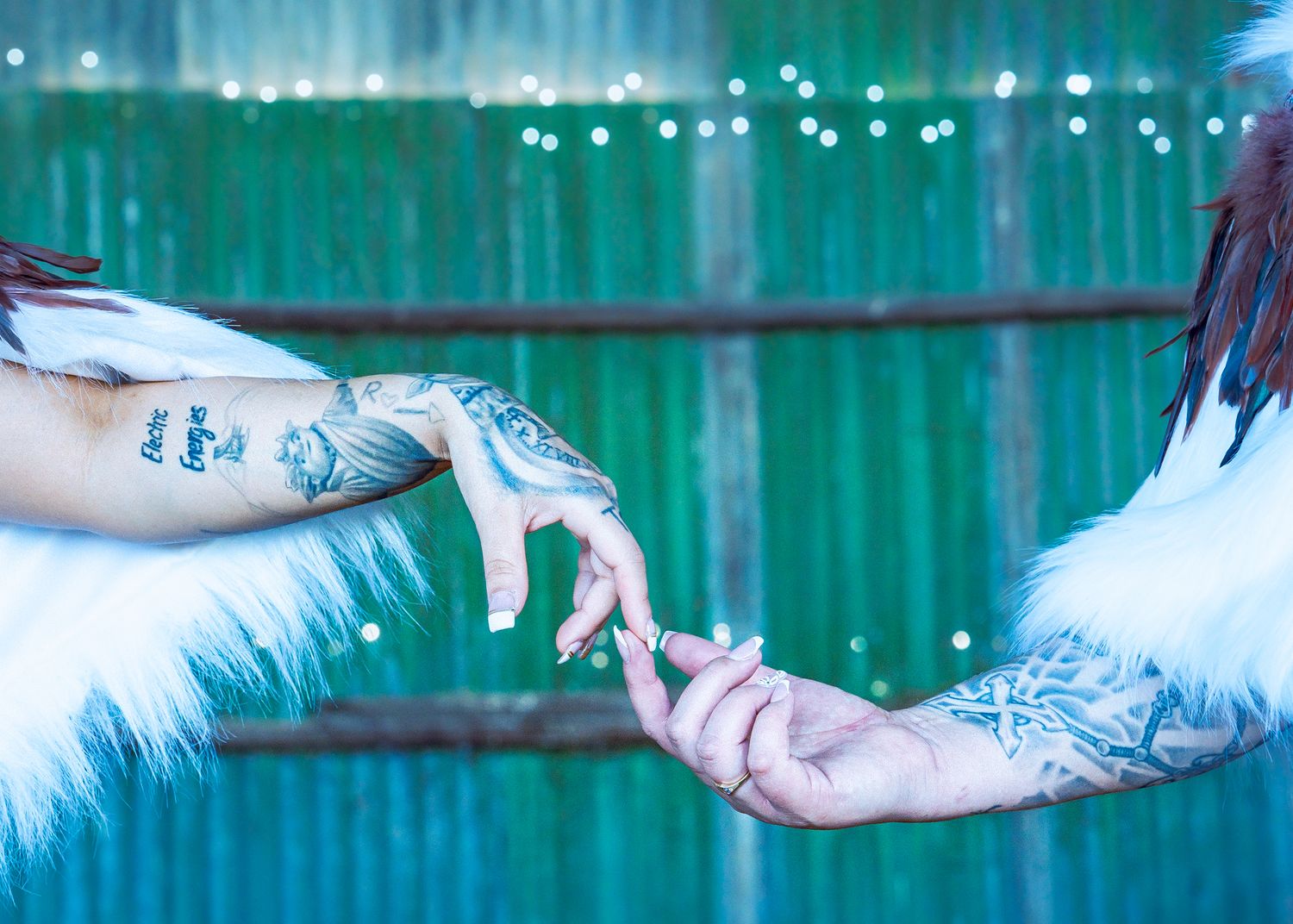 Two tattooed hands reaching towards each other with teal lighting and string lights in the background.