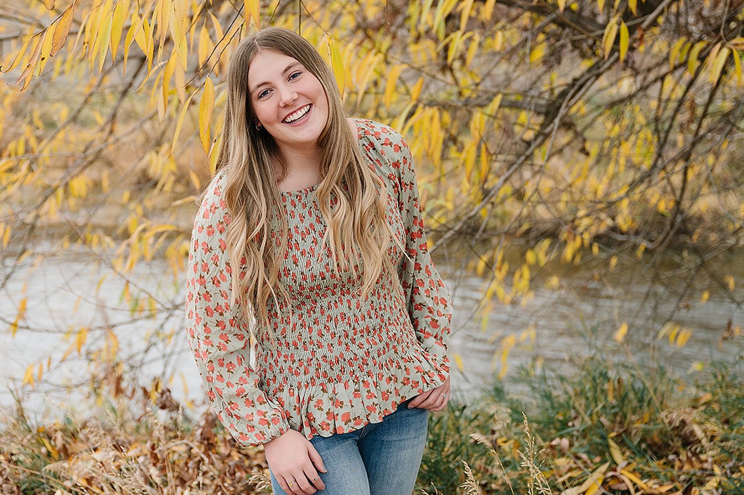 Person in patterned sweater smiles outdoors against autumn foliage background.