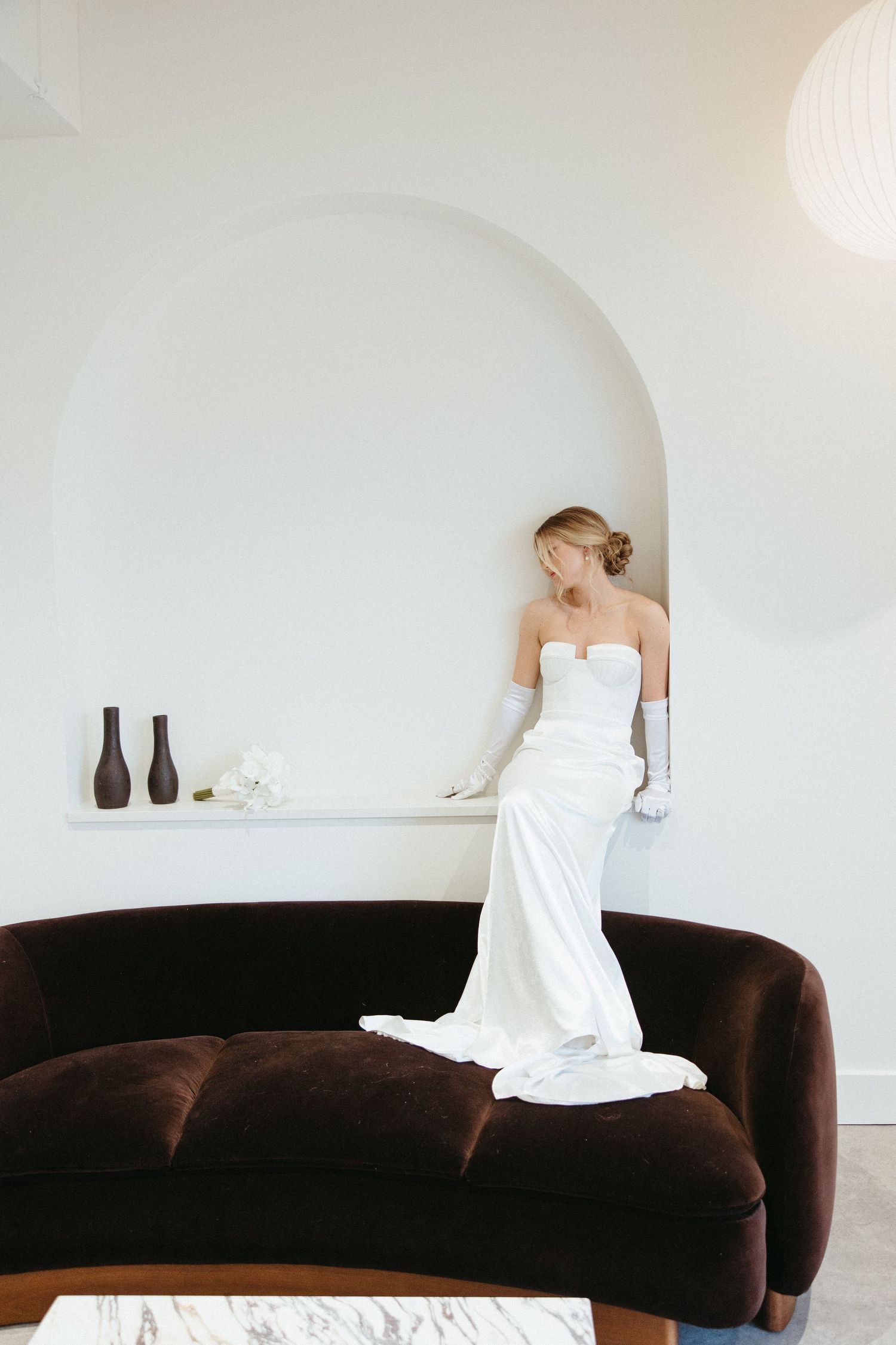 Woman in white dress posing on dark velvet couch in minimalist white room.